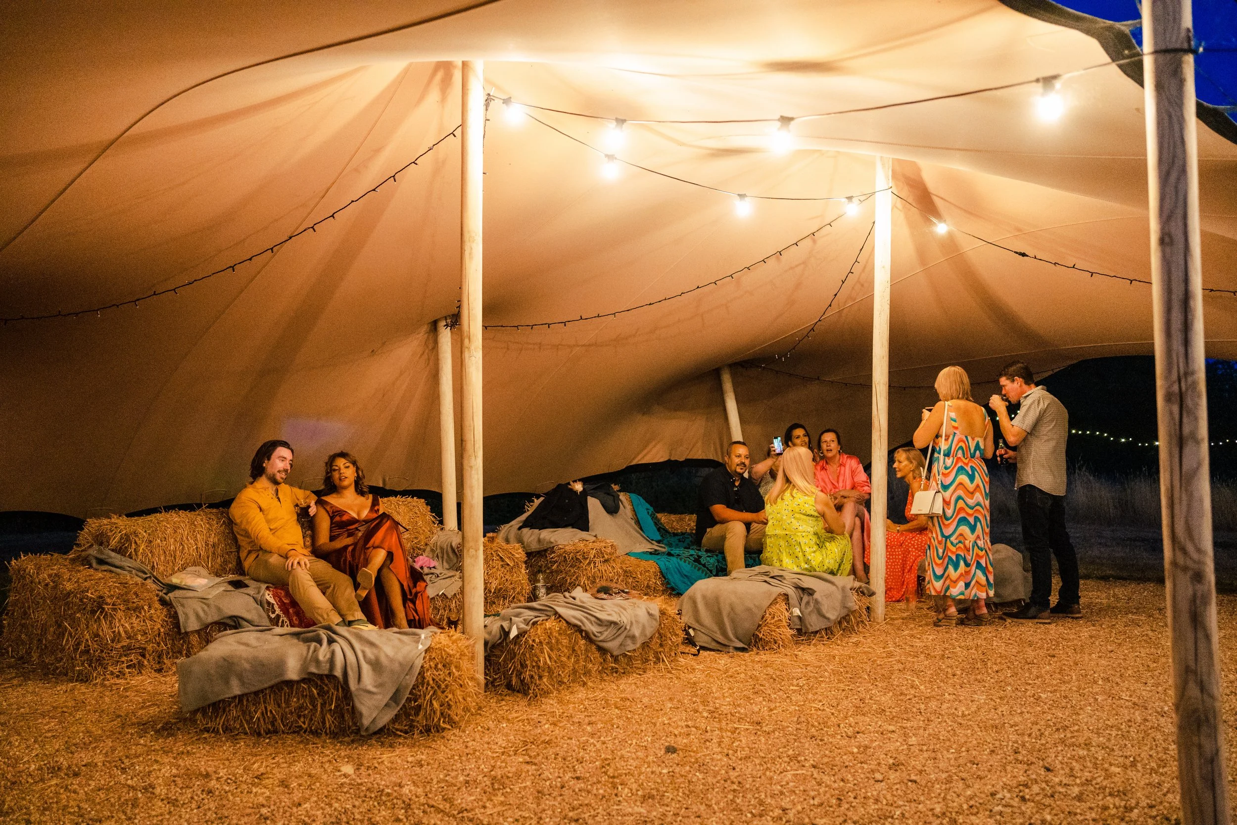 People gathered at an outdoor event under a large beige tent decorated with string lights, sitting on hay bails, with some standing and talking, in a rustic setting during the evening. East Sussex Wedding Farm Glamping