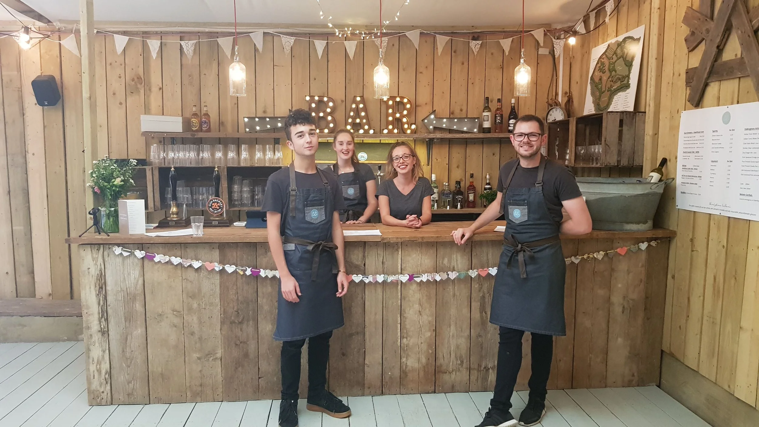 Four waitstaff members standing behind a rustic wooden bar counter with a decorative string of paper hearts. The bar has a large illuminated sign spelling 'BAR' with arrow accents, fairy lights, and various liquor bottles in the background. The staff