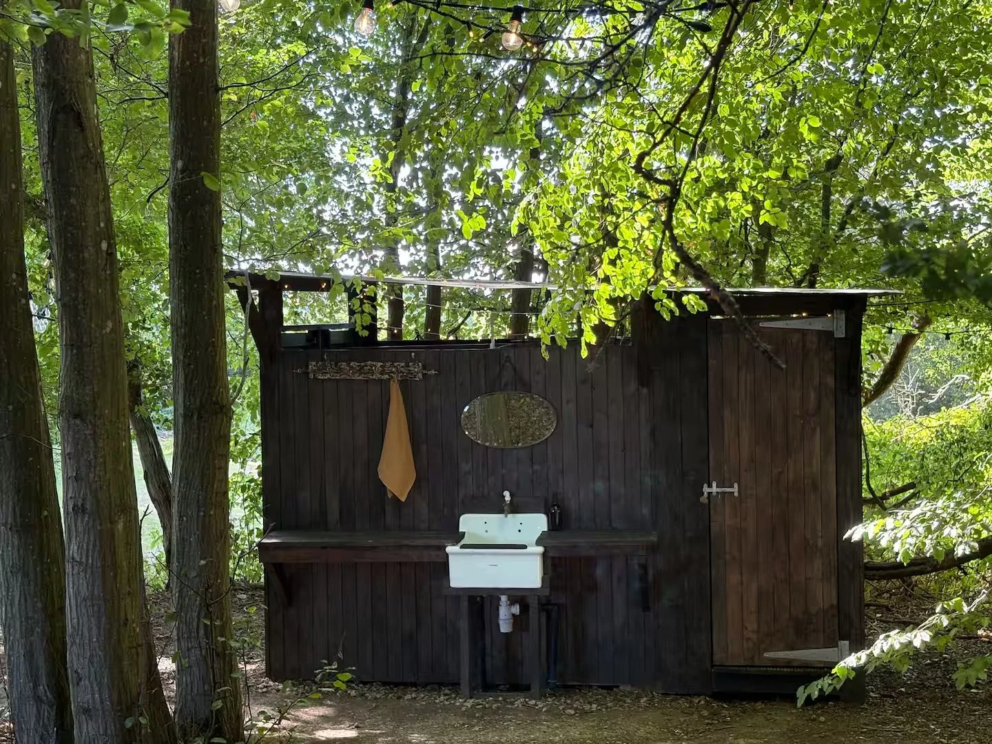 A rustic outdoor sink unit made of dark wood, set against a backdrop of green trees and foliage. The sink is white with a faucet, and a round mirror hangs above it. There is a towel hanging on a hook to the left of the mirror. The structure appears t