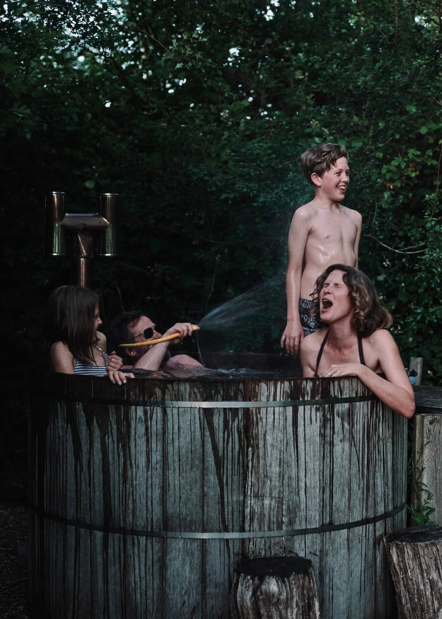People enjoying a hot tub outdoors surrounded by Sussex woodland, with one person spraying water and others laughing.