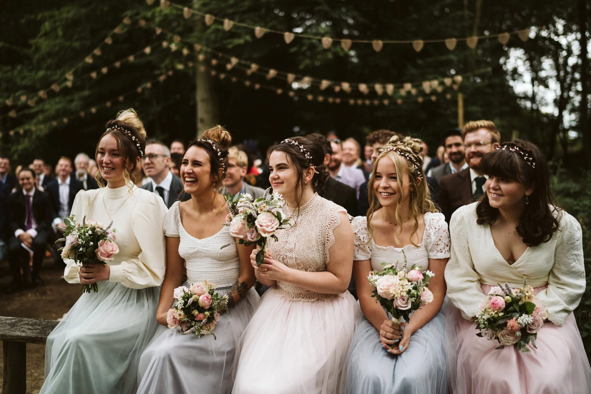 Five women dressed in vintage-style dresses, holding bouquets, sitting outdoors at a wedding ceremony, with an audience of guests behind them, decorated with string lights.