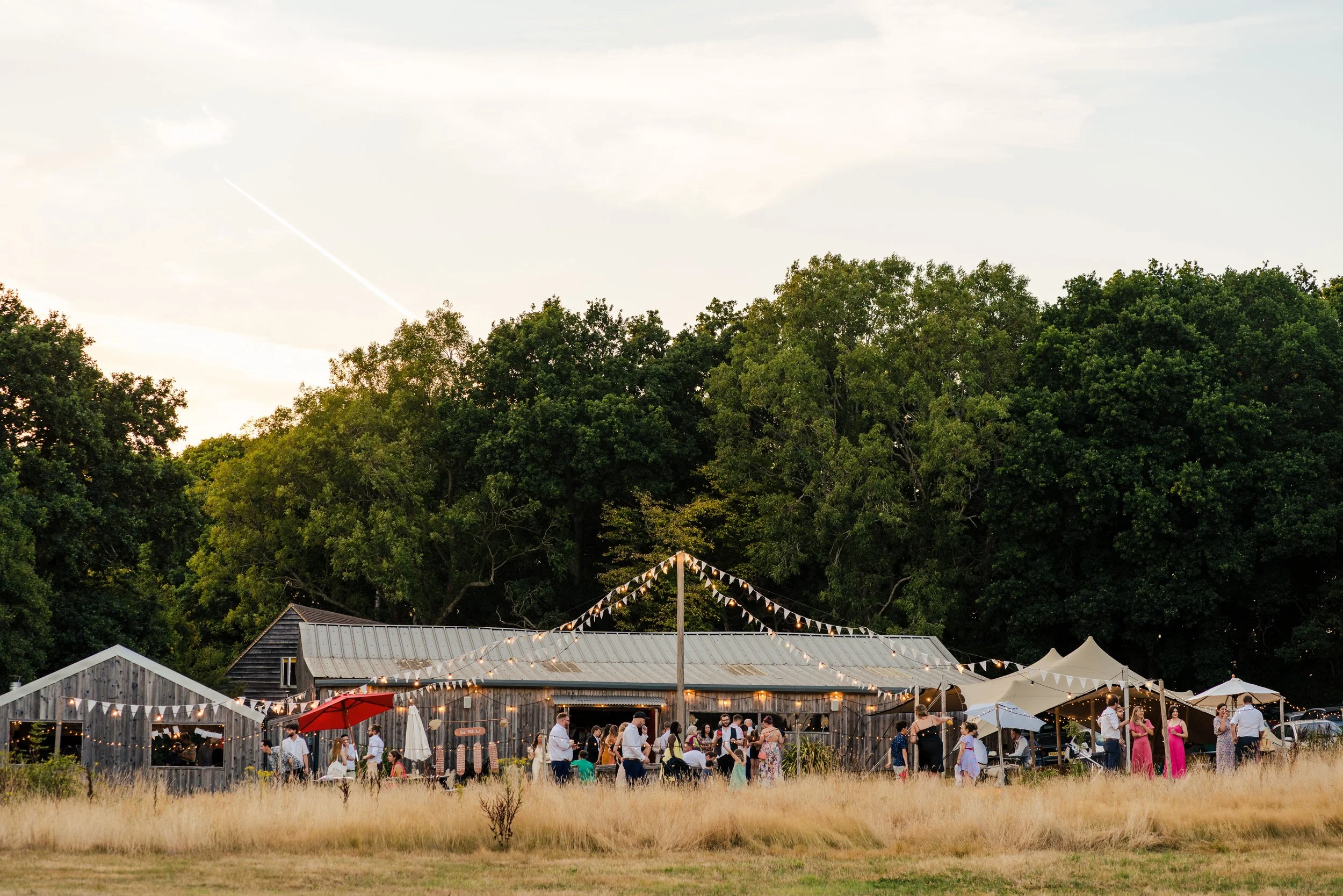 An outdoor wedding reception with guests mingling near rustic wooden buildings, string lights, and tents, set against a backdrop of green trees at sunset. East Sussex Wedding Farm DIY