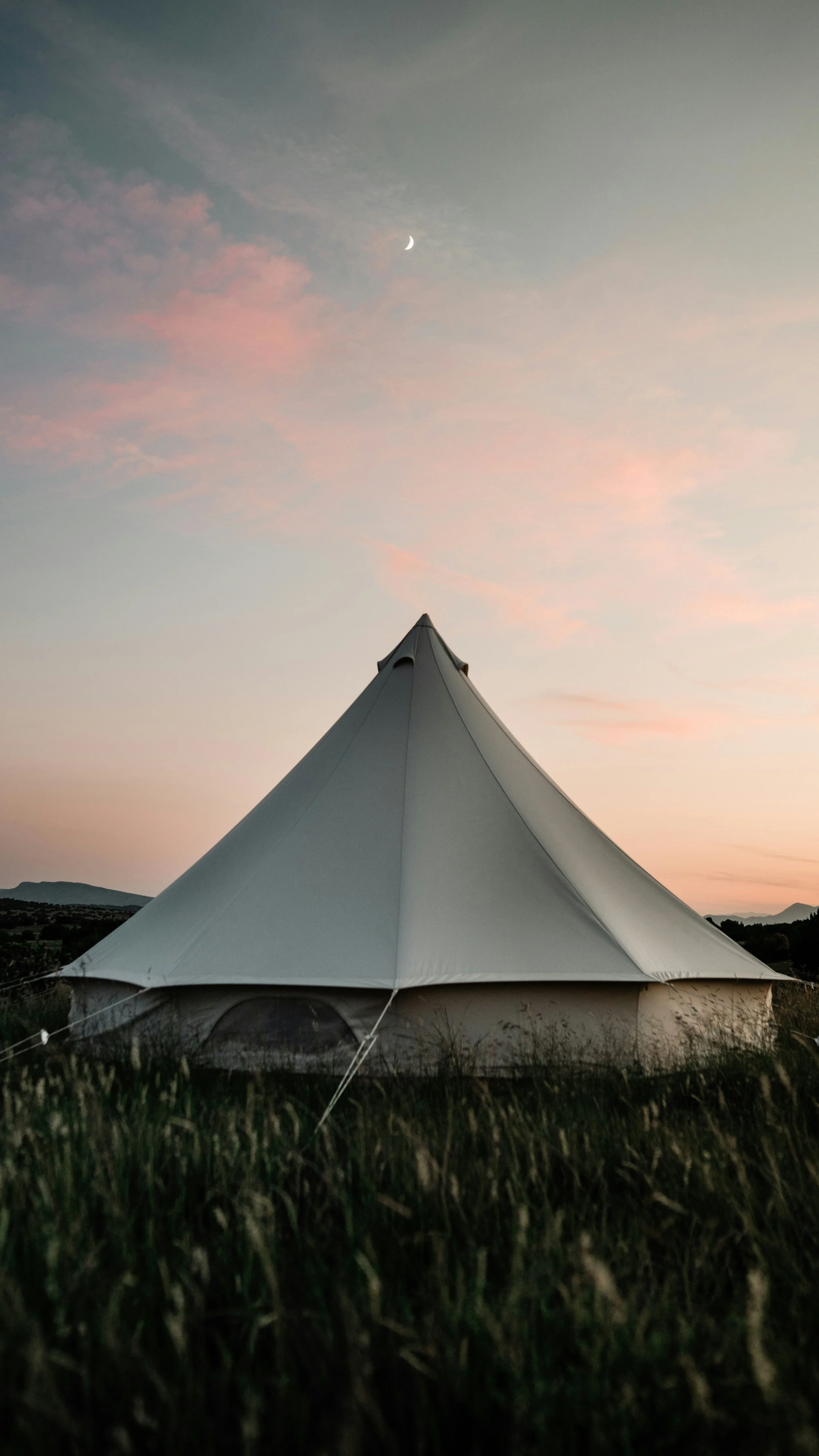 A large white tent in an open field during dusk with a pink and blue sky and a crescent moon overhead. Private Glamping Sussex.