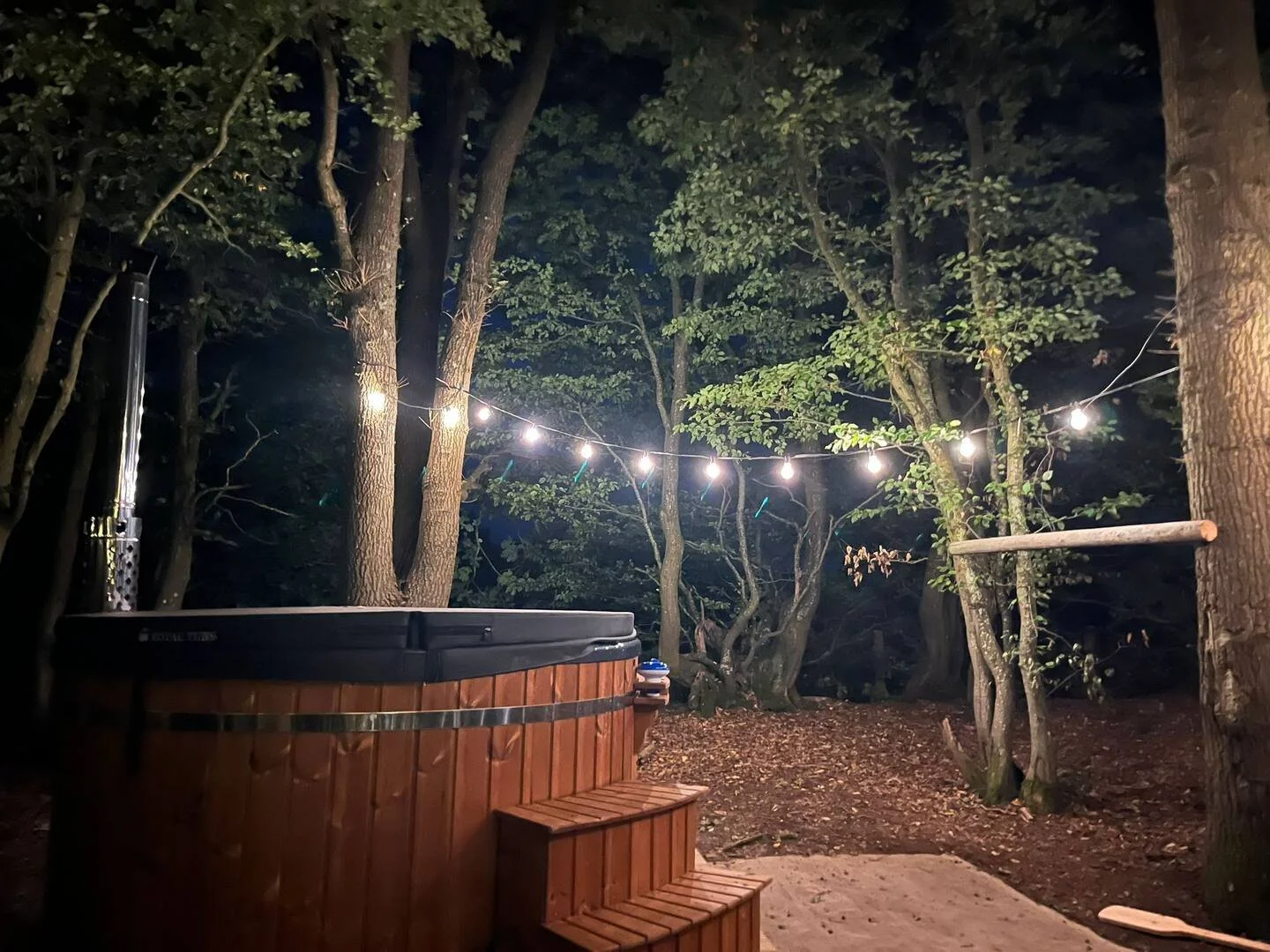 Nighttime outdoor scene with string lights hanging among trees, a hot tub with wooden steps, and a sandy ground area. Private Campsite Glamping Farm.