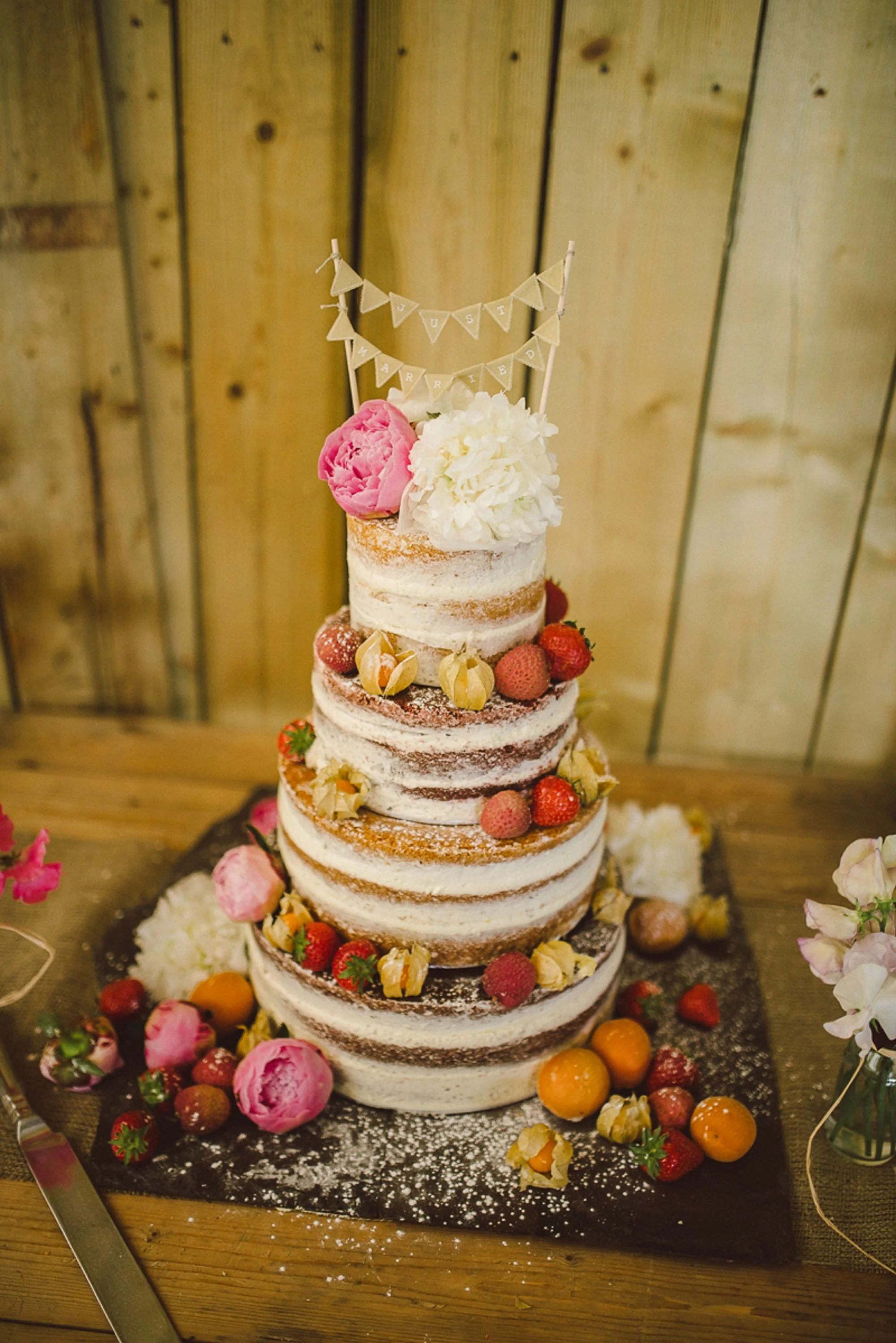 A multi-tiered naked wedding cake decorated with strawberries, physalis, and large white and pink flowers on top. Small pennant banners that read 'Just Married' are on top of the cake.. East Sussex Wedding Glamping Rustic