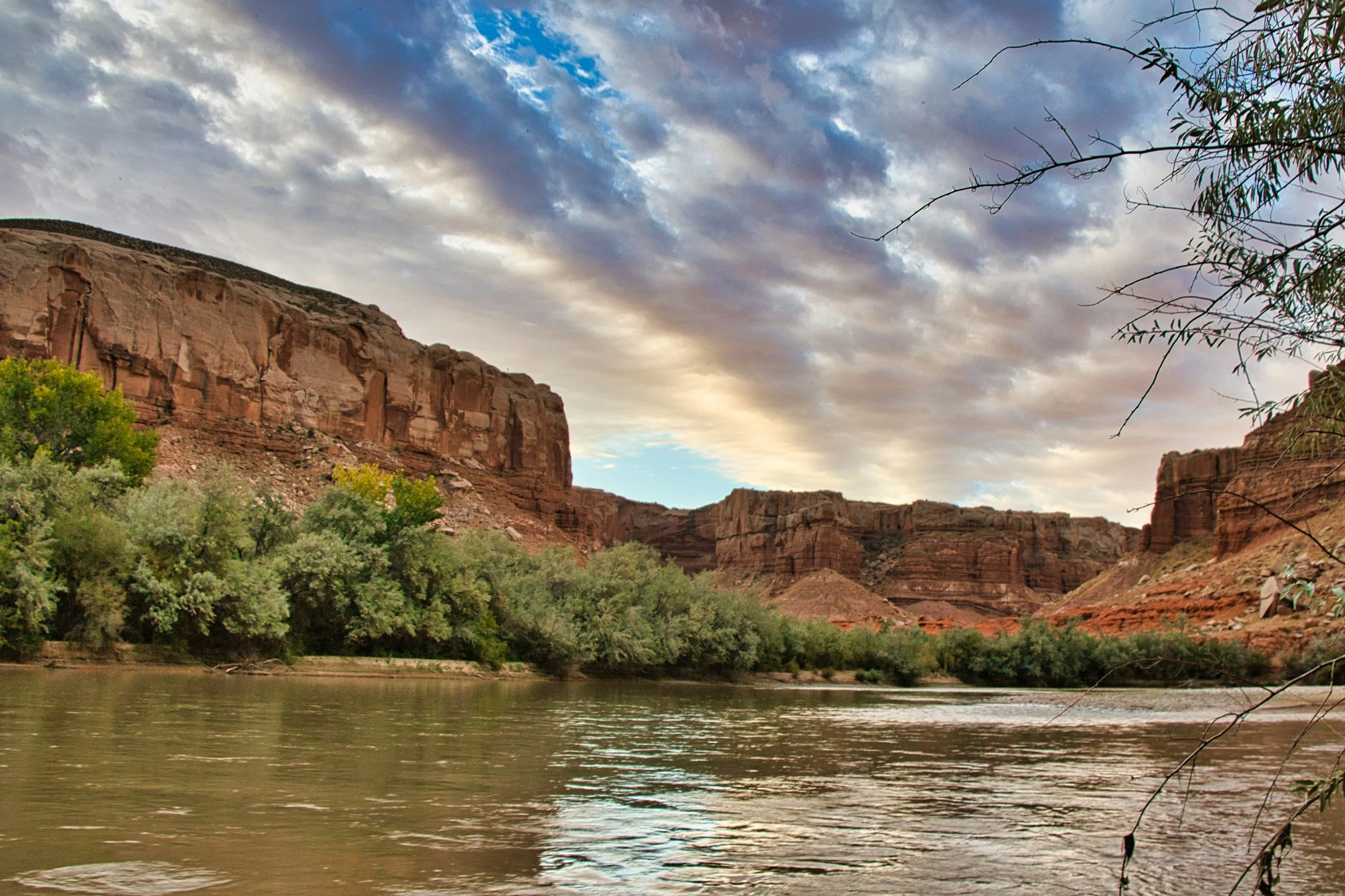 View of a river flowing through a canyon with steep red rock cliffs and green trees along the bank under a cloudy sky.