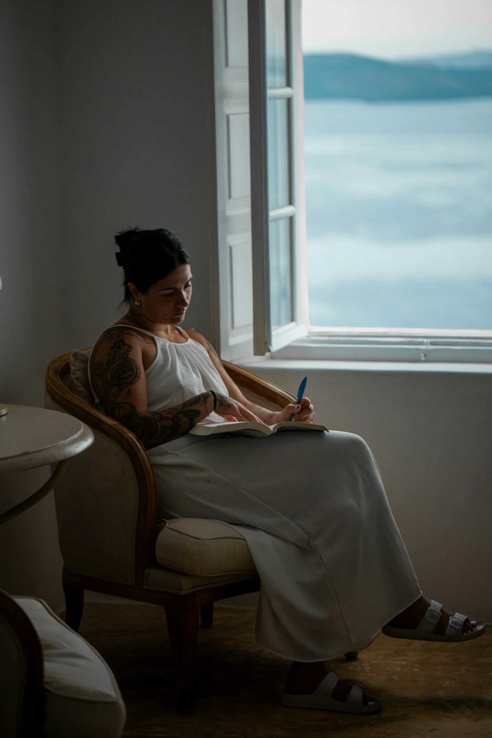 A woman sitting on a chair near a window, looking at her phone with a book on her lap, and the ocean visible outside.