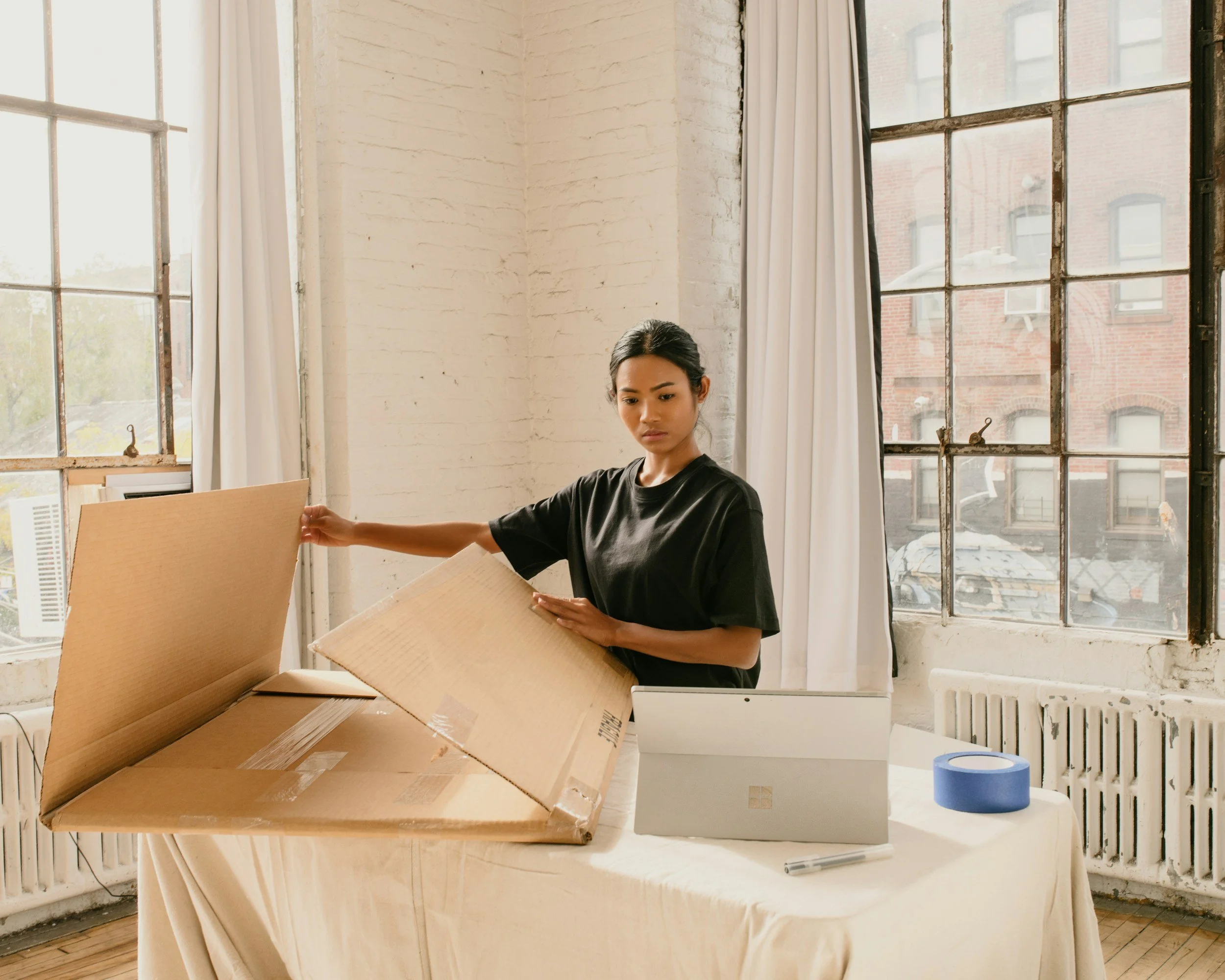 A woman with dark hair standing in a bright room with large industrial-style windows, holding a large cardboard box, with more boxes and packing tape on a table nearby.