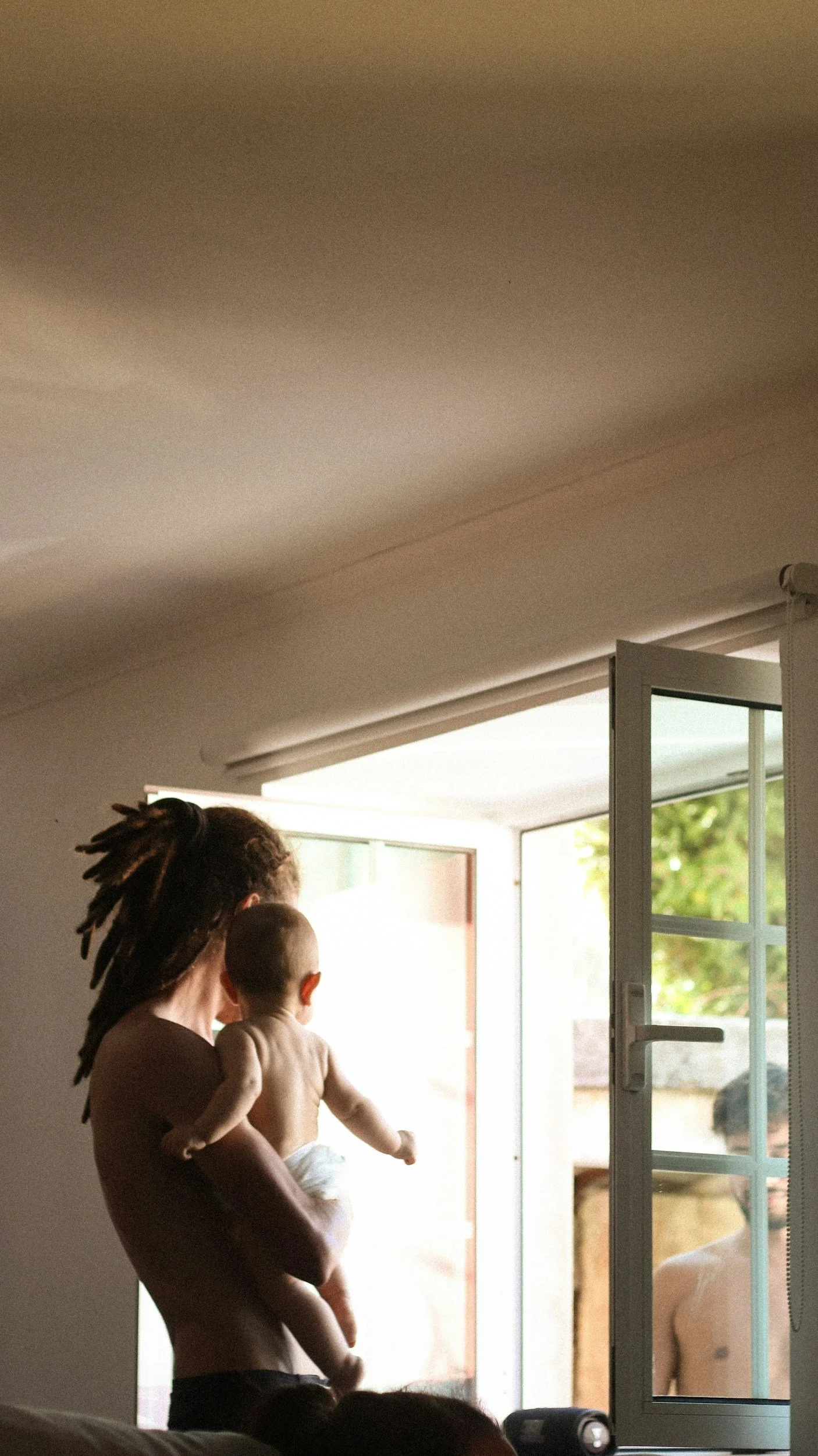A person with dreadlocks holding a baby near an open door, with sunlight coming in and greenery outside.
