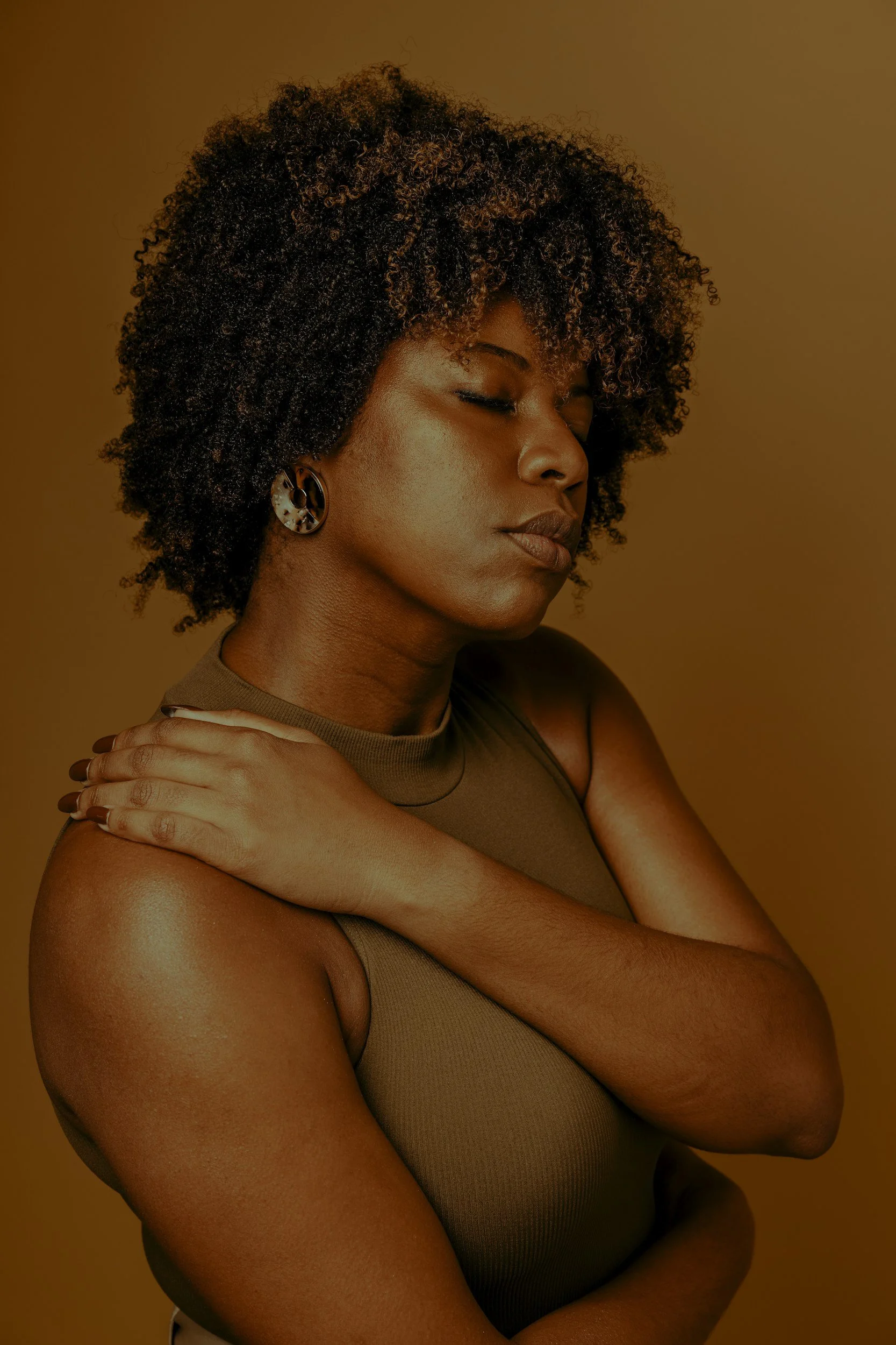 A woman with voluminous curly hair, closed eyes, and earrings, embracing herself against a warm brown background.