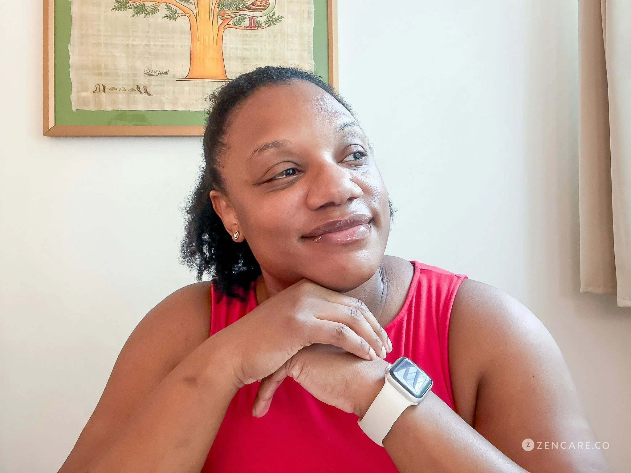 A woman with dark, curly hair, wearing a pink sleeveless top and a smartwatch on her left wrist, is smiling softly and resting her chin on her right hand in a room with light-colored walls, a window with a curtain, and a framed picture of a tree on the wall behind her.