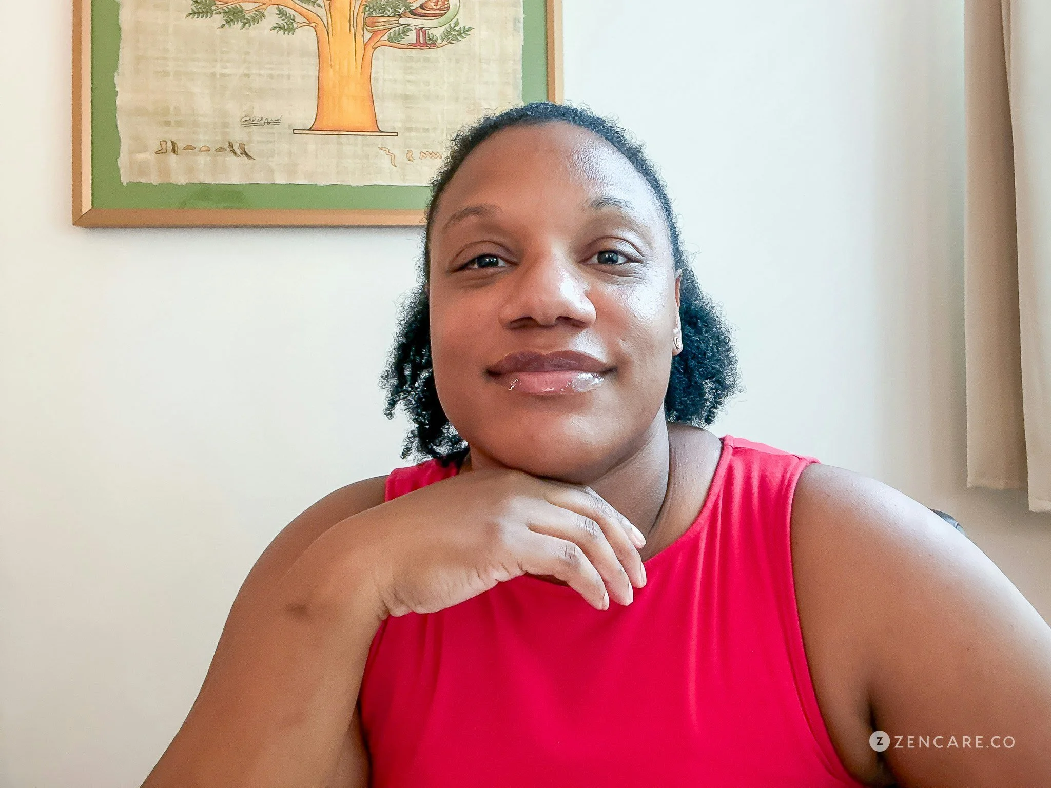 A woman with dark curly hair and a bright pink sleeveless top is taking a selfie in front of a light-colored wall. Behind her, there is a framed illustration of a tree with branches, leaves, and some writing in a different language.