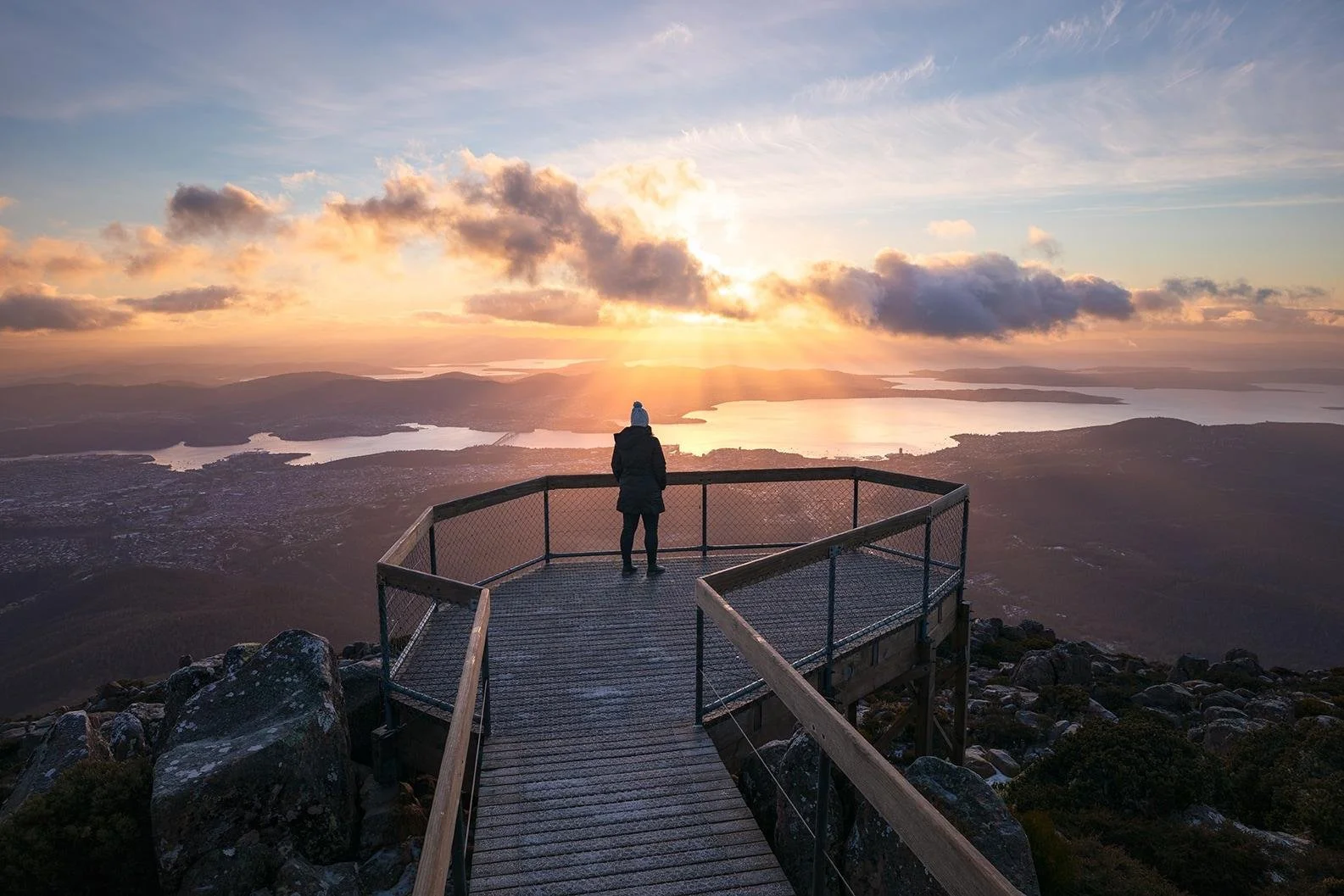 A person on a viewing platform on a mountain at sunset, overlooking a landscape of lakes, hills, and a partly cloudy sky.