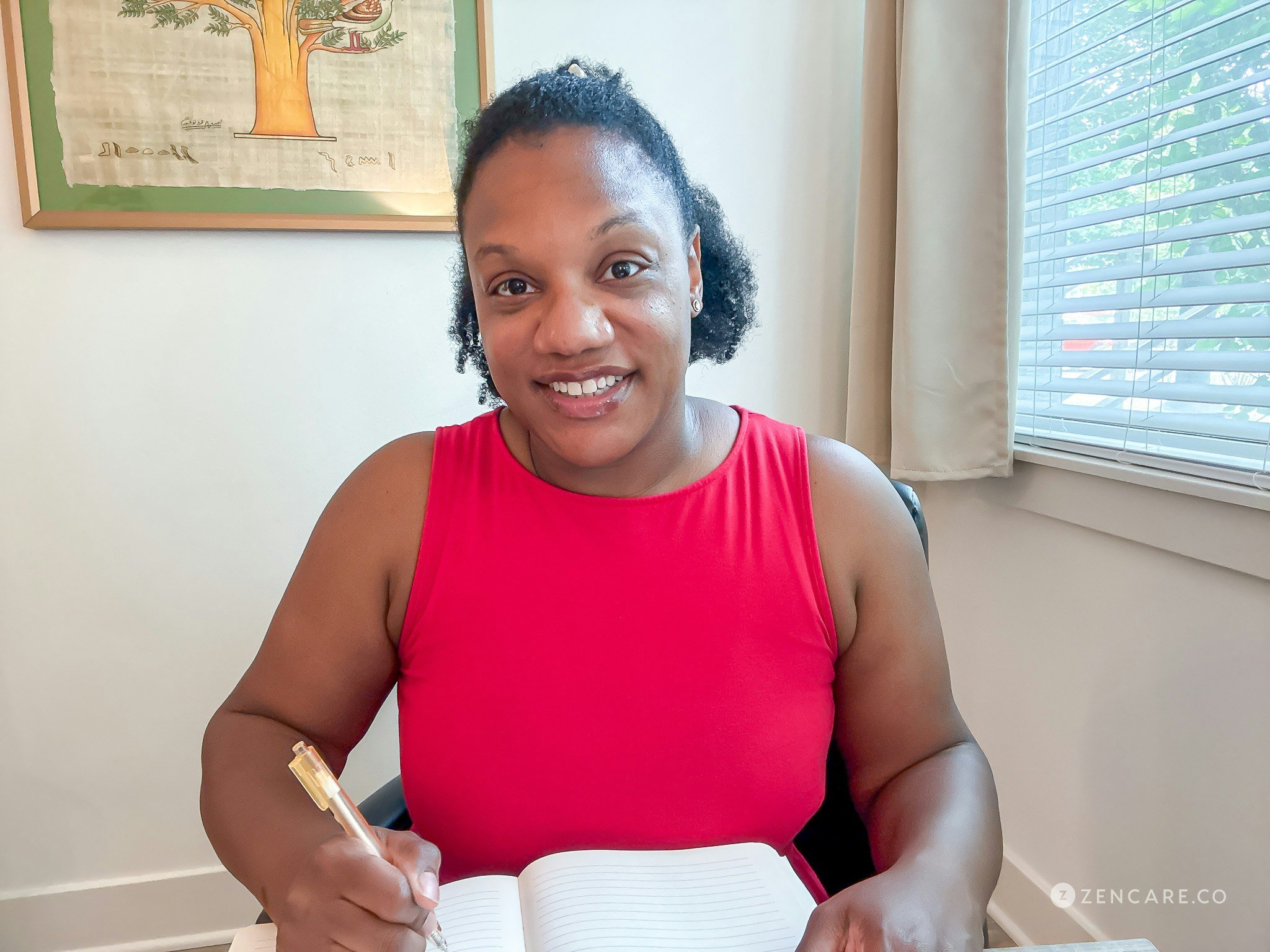 A woman with dark curly hair wearing a red sleeveless top, sitting at a table, writing in a notebook with a pen, in a room with a window, white walls, a zebra curtain, and a painting of a tree on the wall.