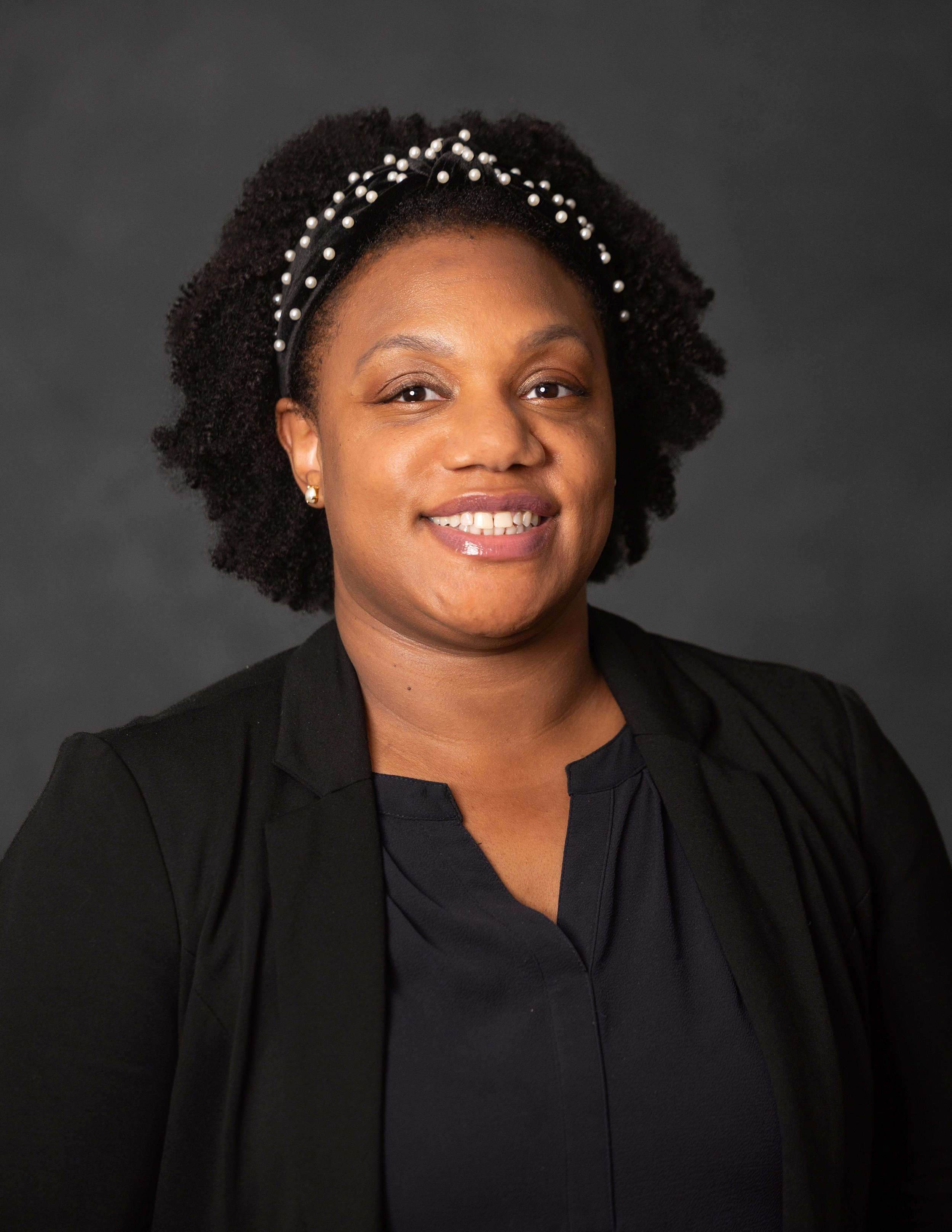 A woman with short, curly hair wearing a black blazer, black top, pearl earrings, and a pearl headband, smiling against a dark gray background.