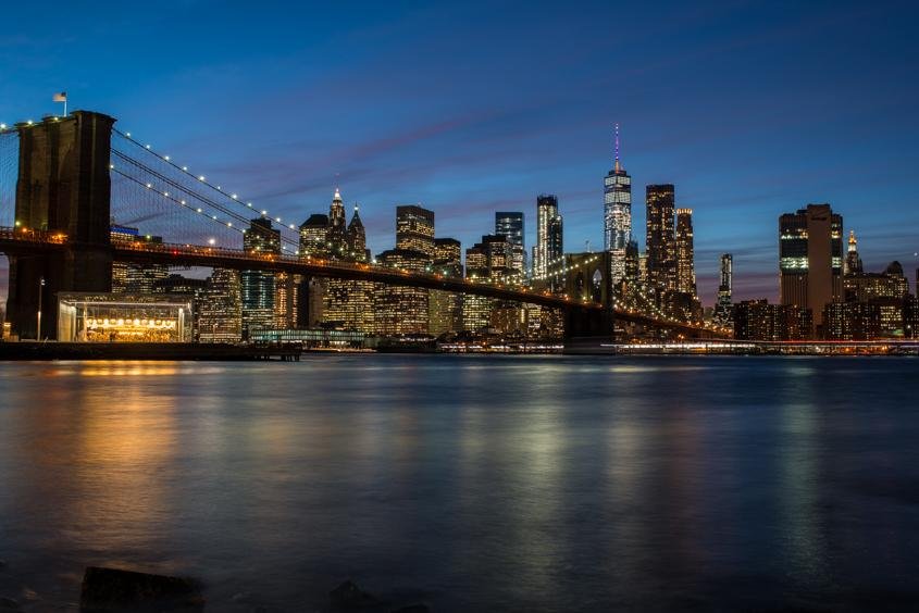 Nighttime view of the New York City skyline with the Brooklyn Bridge in the foreground, illuminated skyscrapers, and reflections on the river.