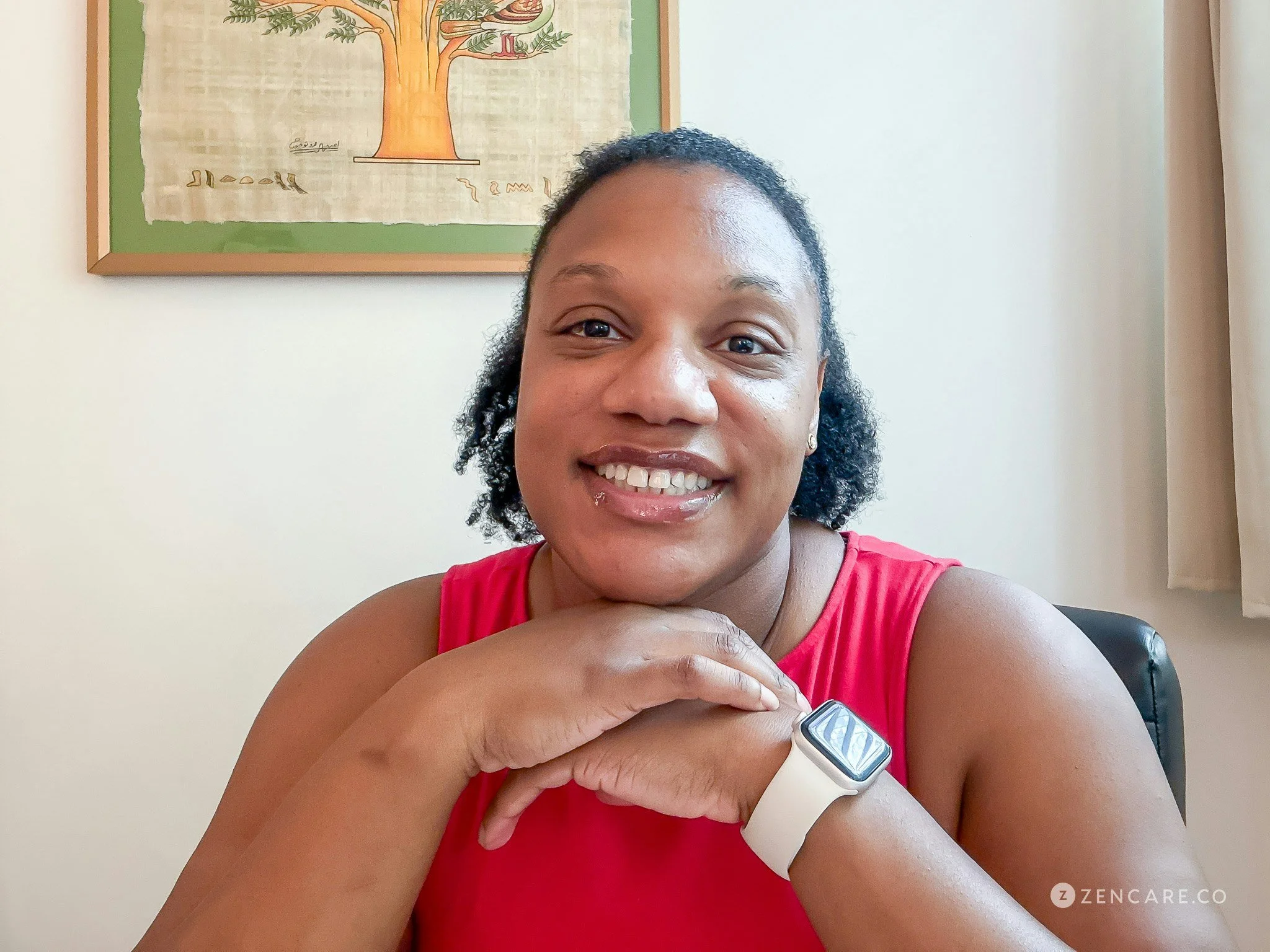 A woman with curly hair and a bright smile taking a selfie indoors. She is wearing a pink tank top, a smartwatch on her left wrist, and appears to be holding an object near her chest. In the background, there is a framed artwork of a tree on a beige wall and a curtain on the right side.