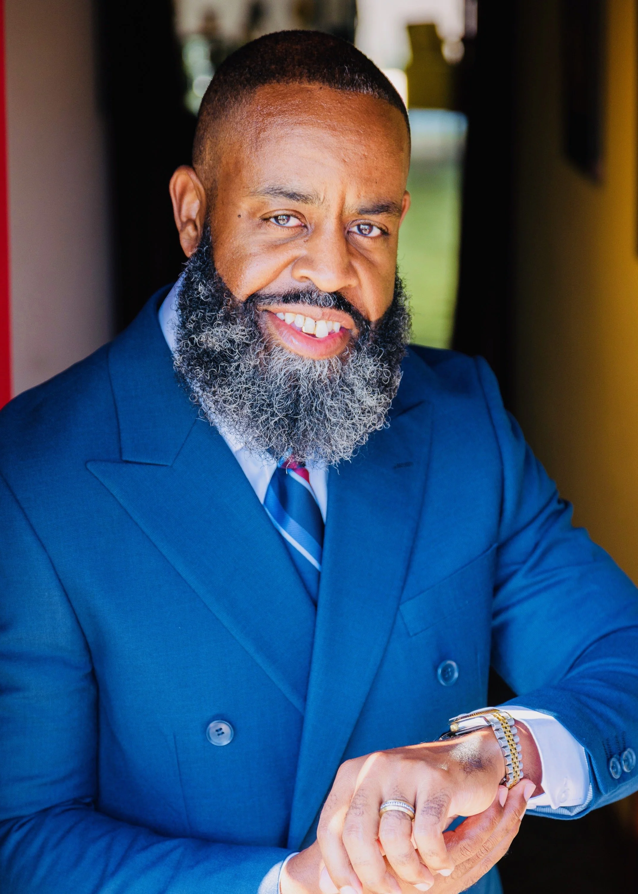A man in a blue suit with a white shirt and striped tie, smiling and looking at the camera while adjusting his watch. He has a thick beard with some gray, short black hair, and is wearing rings and a gold bracelet.