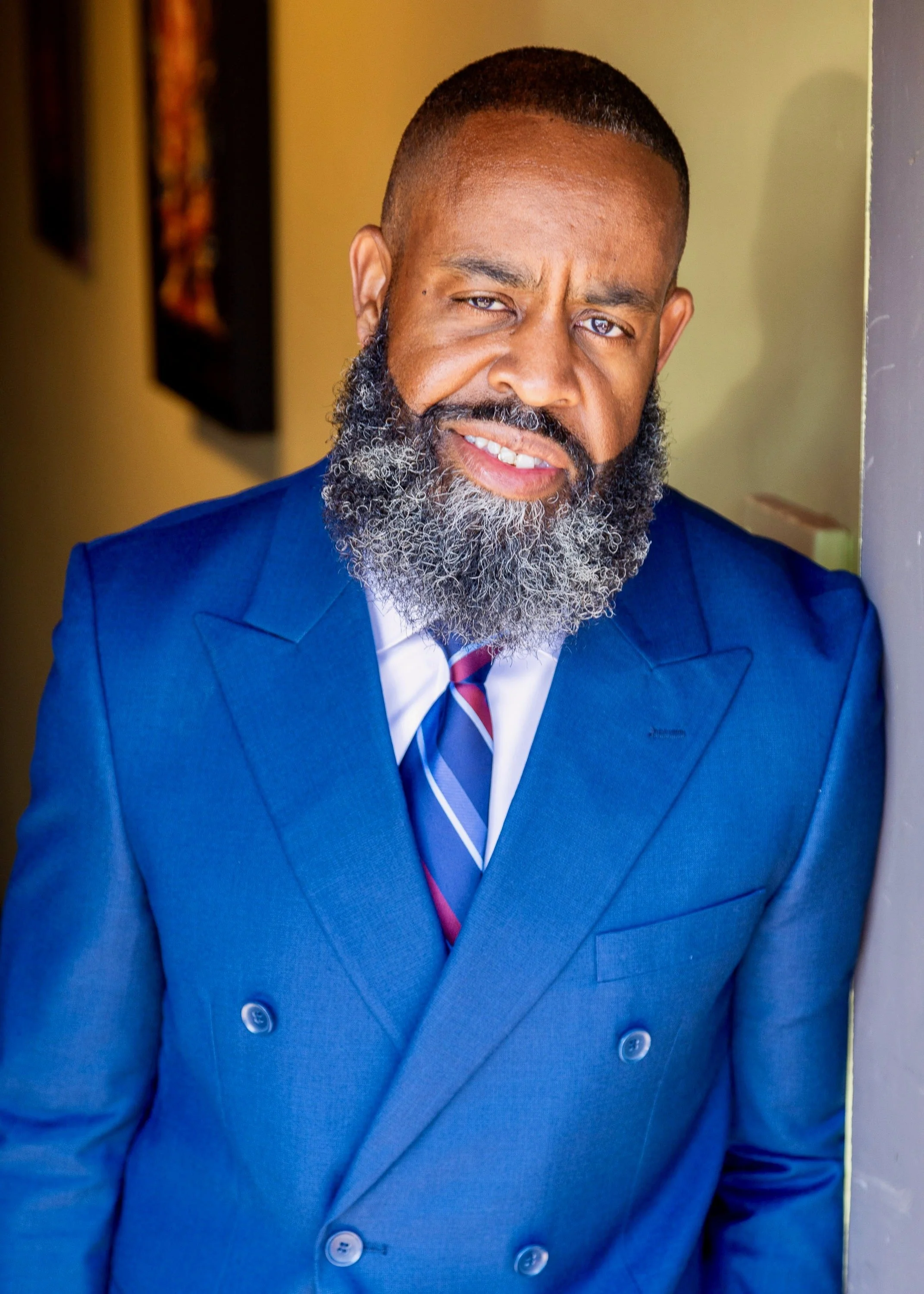 A man with a beard and short hair wearing a blue double-breasted suit, white shirt, and striped tie, leaning against a wall with a neutral expression.