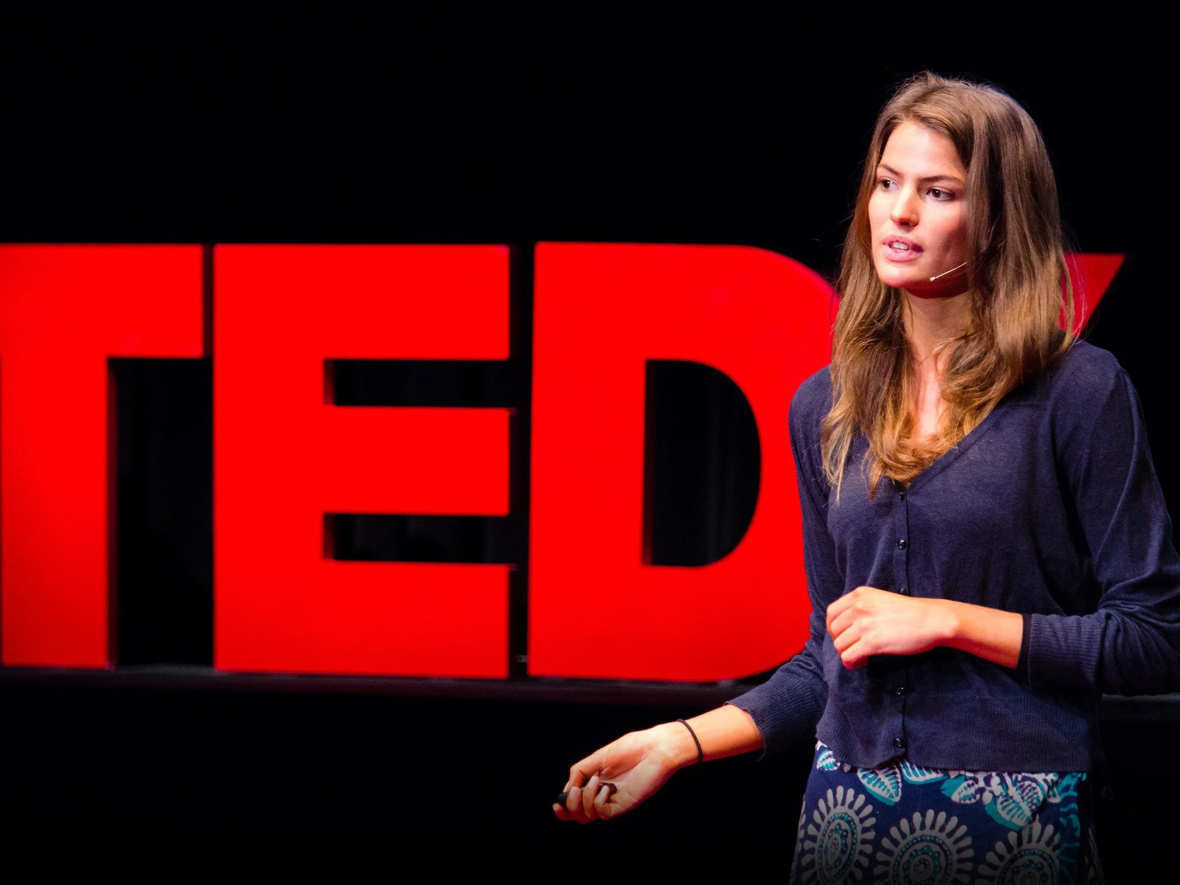 A woman giving a presentation on stage, standing in front of a large red and black "TED" sign.