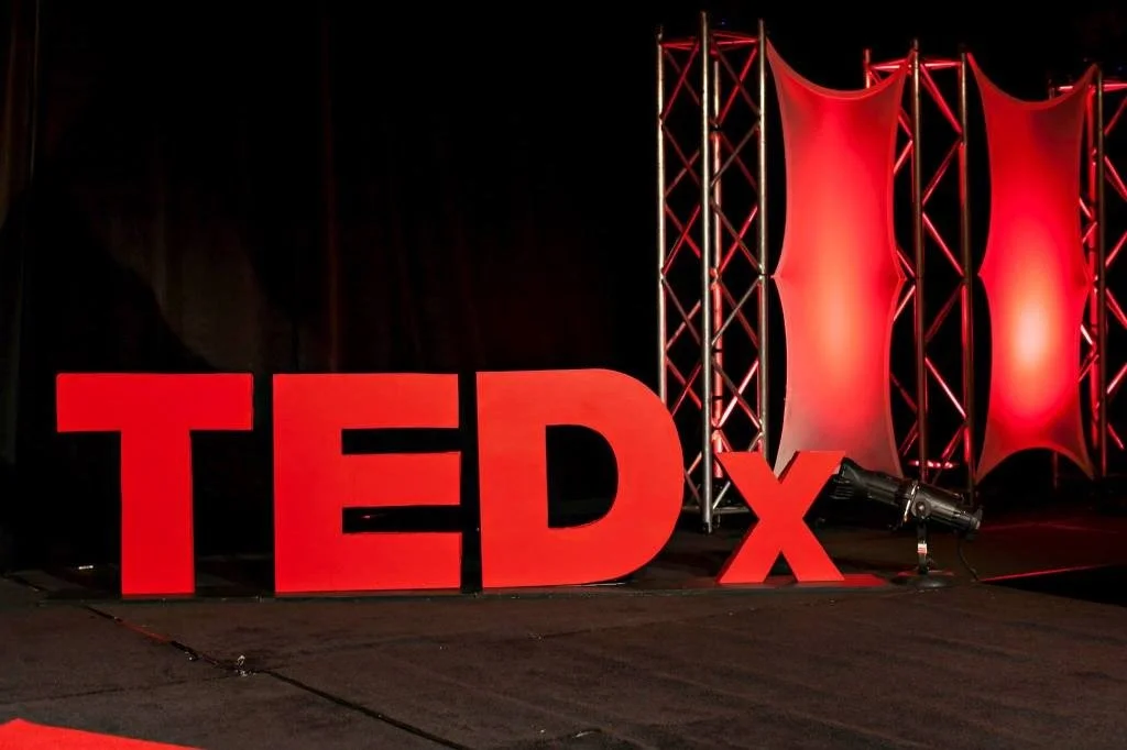 Stage setup with large red TEDx letters and red fabric panels, illuminated by stage lighting.