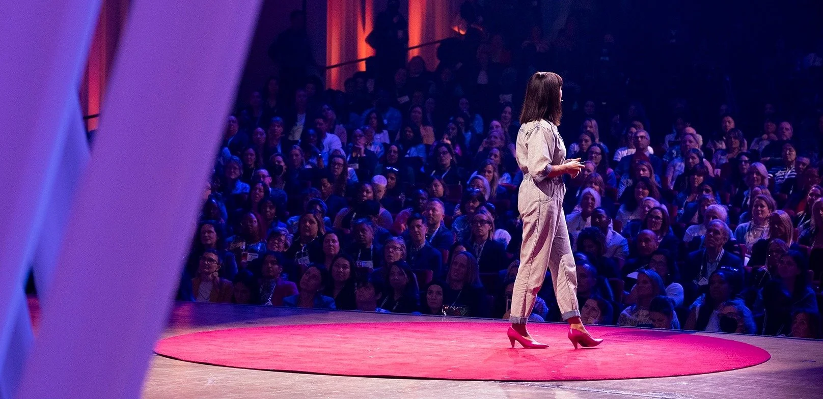 A woman wearing beige pants and a blouse speaking on stage at a conference. The audience is sitting and watching attentively in the background.