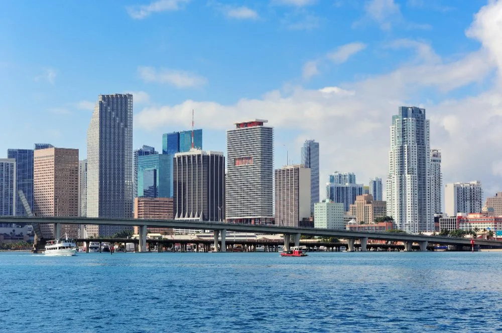 City skyline with tall skyscrapers by the water, featuring a bridge and boats in the foreground, under a partly cloudy sky.