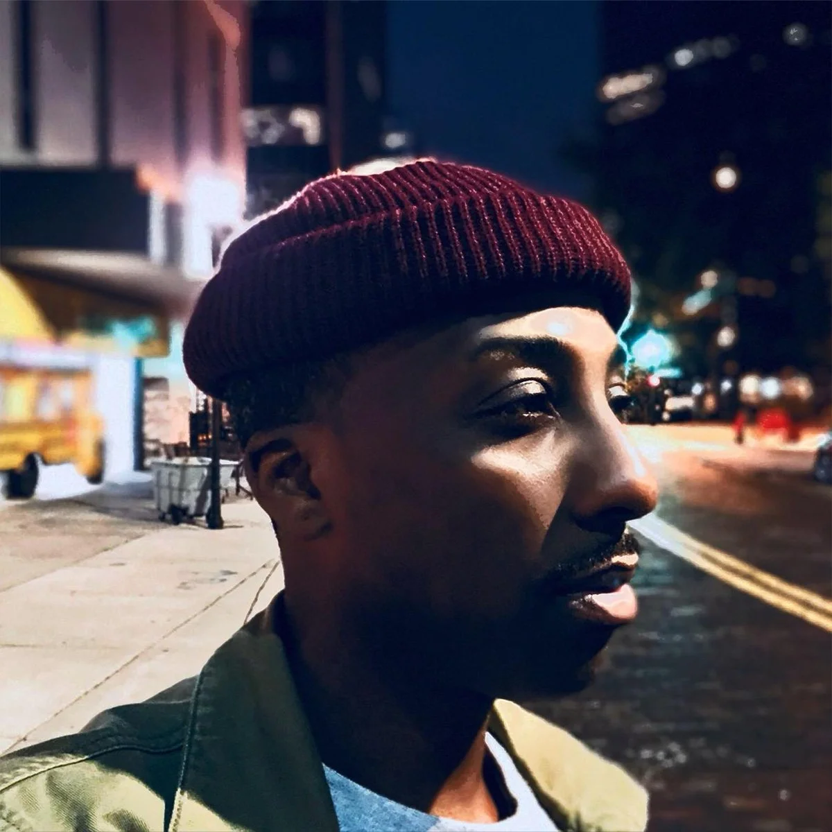 A young man wearing a red knit beanie, standing on a city sidewalk at night, with streetlights and blurred buildings in the background.