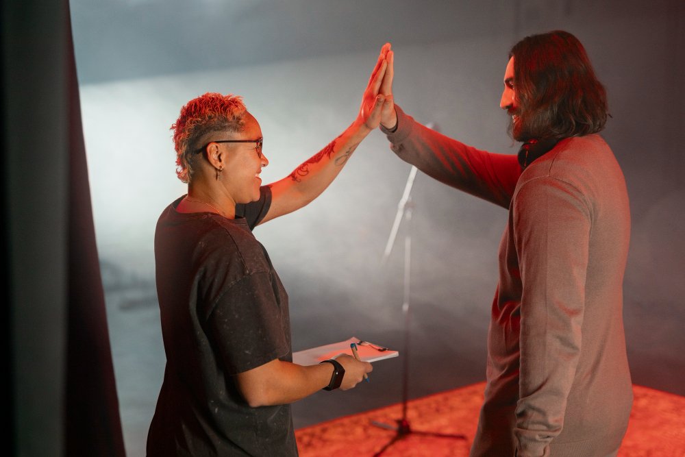 Two people giving a high-five on a stage with a foggy background, one holding a notebook, in a setting with stage lighting.