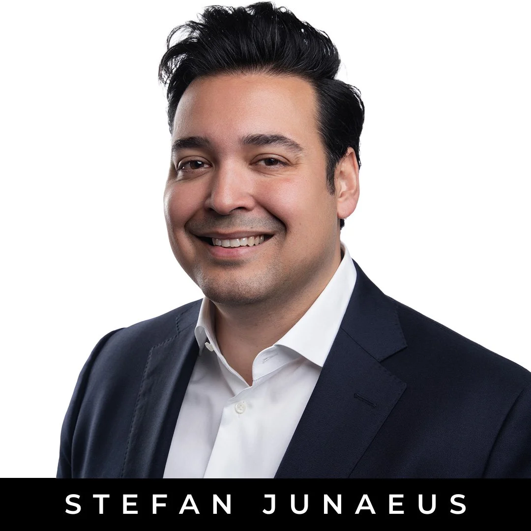 Headshot of Stefan Junaeus smiling, wearing a white shirt and dark suit against a white background.