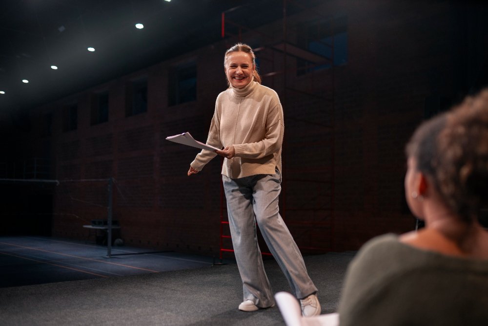 A woman on stage smiling and holding papers, engaging with an audience member in a dark indoor setting.