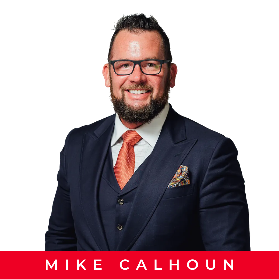 A man with glasses and a beard wearing a dark suit, white shirt, and salmon-colored tie, smiling against a plain white background, with a name tag that reads 'Mike Calhoun' in white letters on a red band at the bottom.