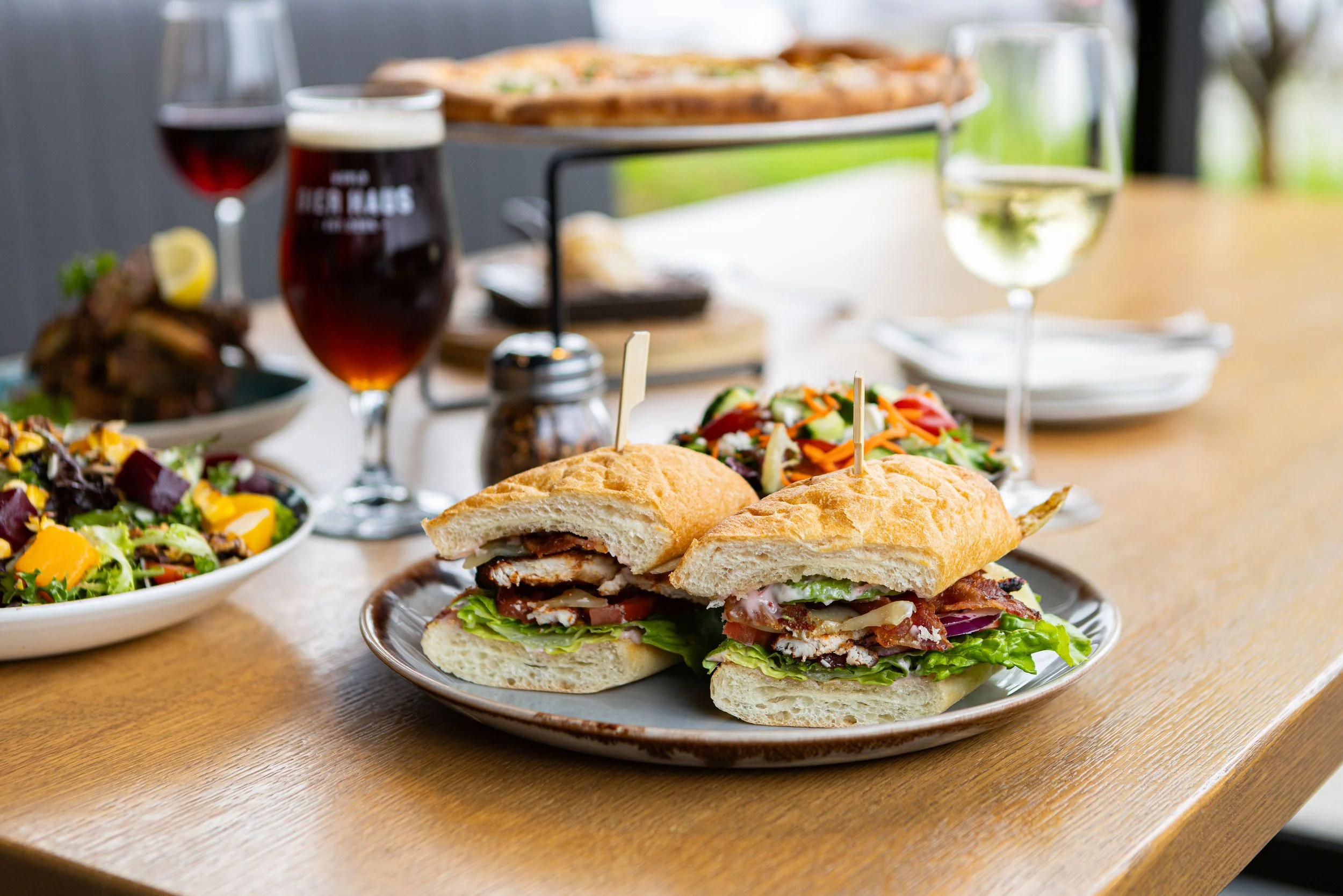 Plate with a cut sandwich containing lettuce, tomato, cheese, and meat on a wooden table, with salad, beer, wine, and other dishes in the background.