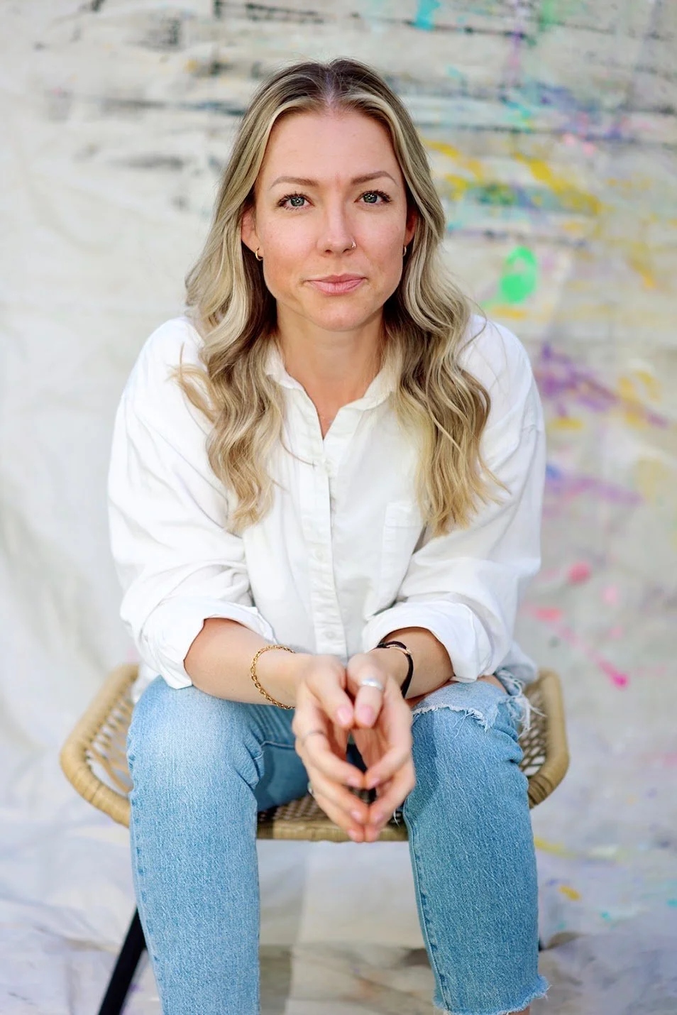 A woman with blonde hair, wearing a white shirt and ripped blue jeans, sitting on a wicker chair with her hands clasped, against a colorful, abstract background.