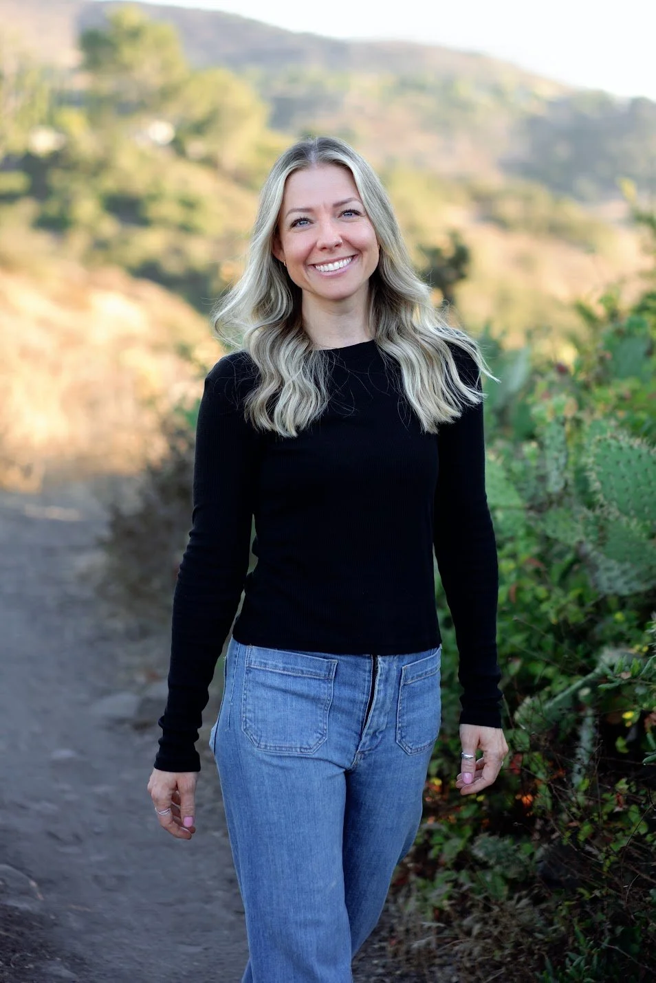 A woman with long blonde hair, wearing a black long sleeve shirt and blue jeans, smiling while walking outdoors on a trail with green hills and vegetation in the background.