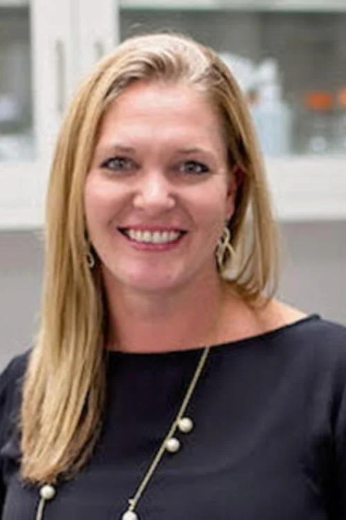Portrait of a smiling woman with blonde hair, wearing a black top and a pearl necklace, standing indoors.