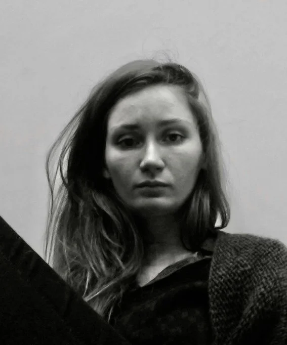 Black and white photo of a young woman with long wavy hair, looking serious, sitting against a plain wall.