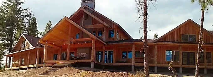 A large , newly built home with log pillars running along a wrap around deck. The home is a natural pine color and is centered in front of a gray sky, pine trees to the left of the home.