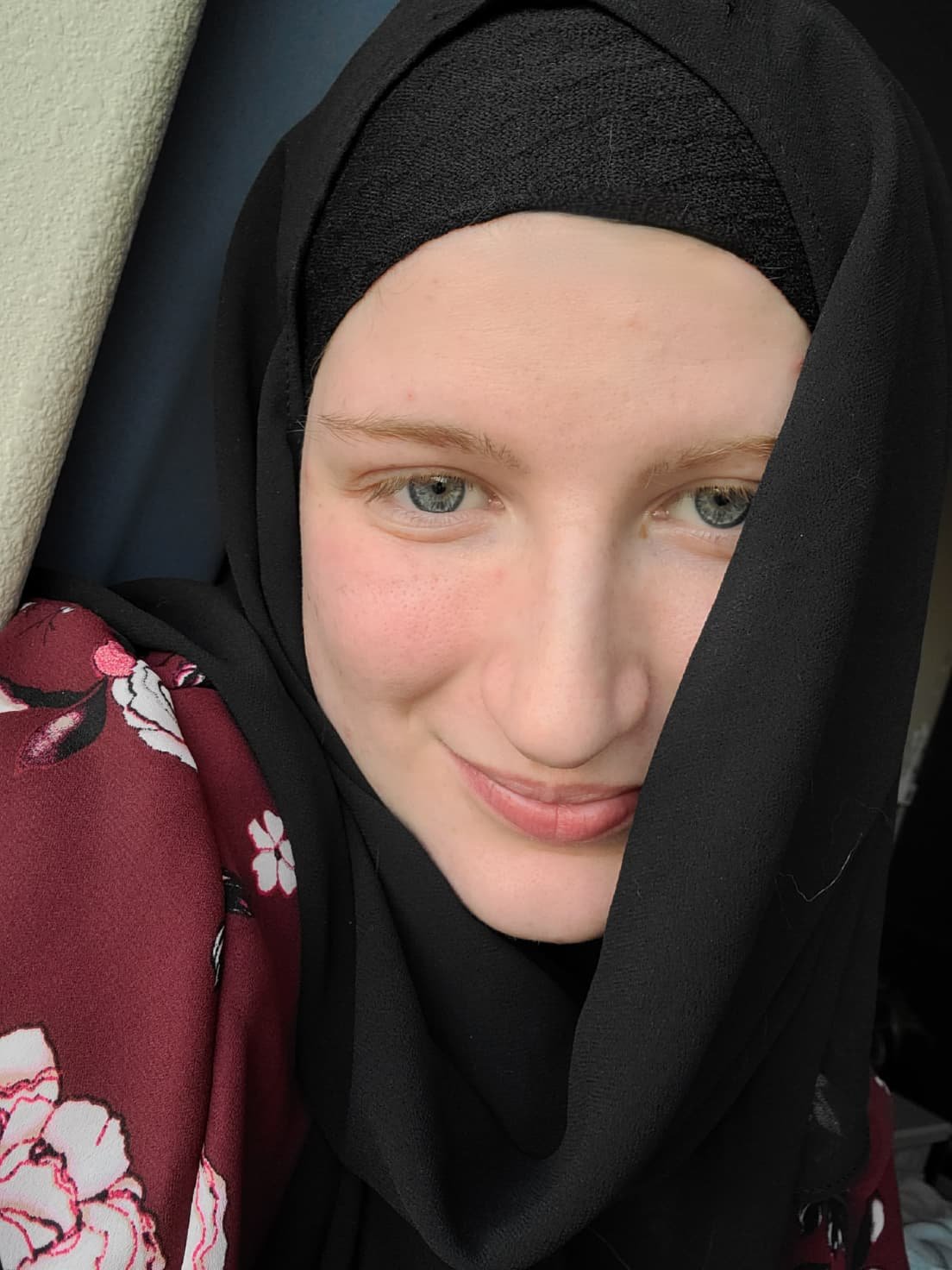 Blue eyed white woman wearing a red floral shirt and a black hijab, slightly covering her right cheek.