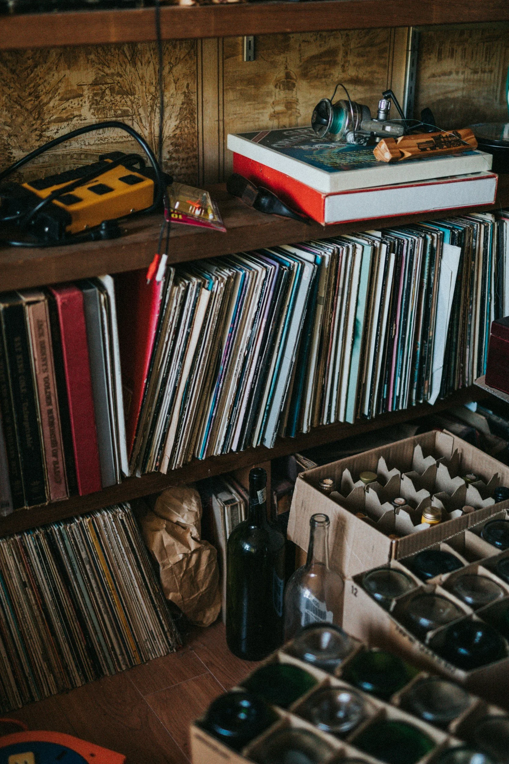 A wooden shelf filled with vinyl records, a cardboard box holding bottles, and various small objects including a yellow device and headphones.