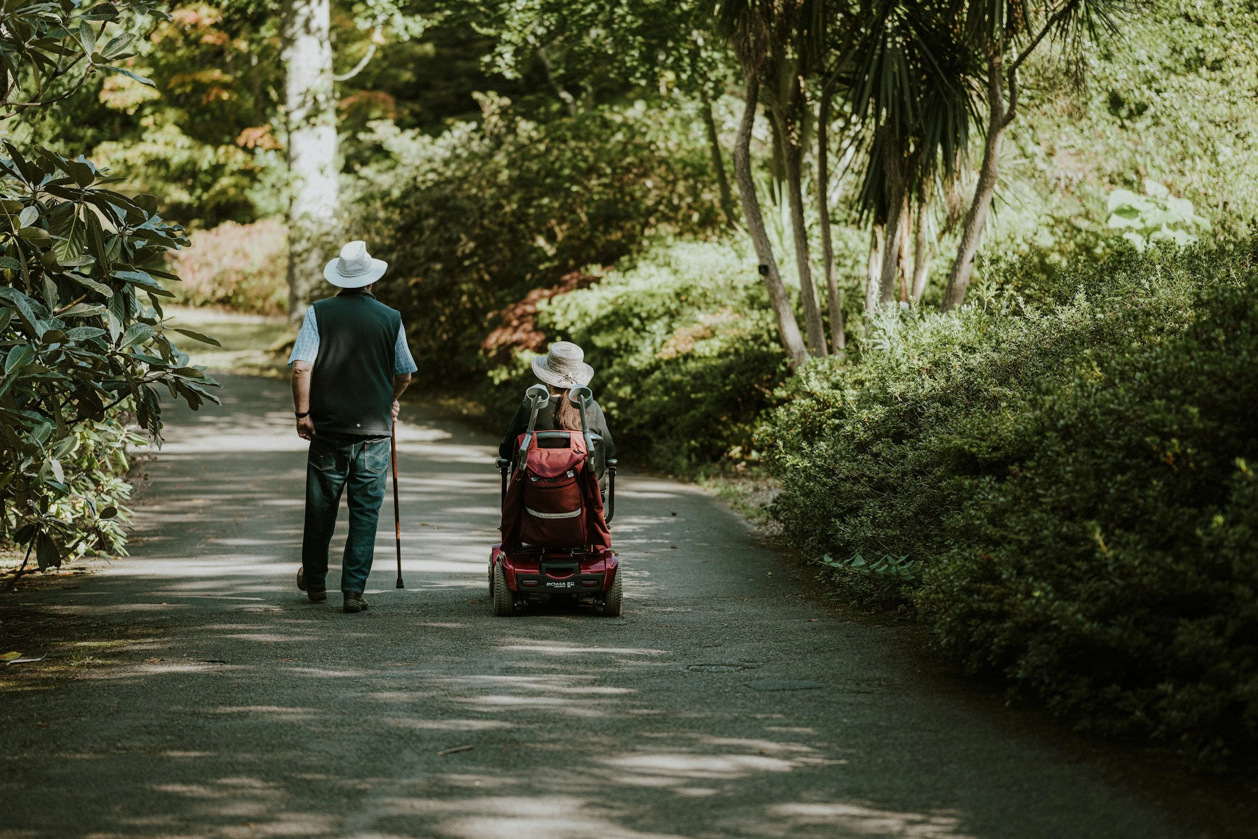 An elderly person with a mobility scooter beside a person walking with a cane, both wearing wide-brimmed hats, walking on a shaded, tree-lined path in a park.