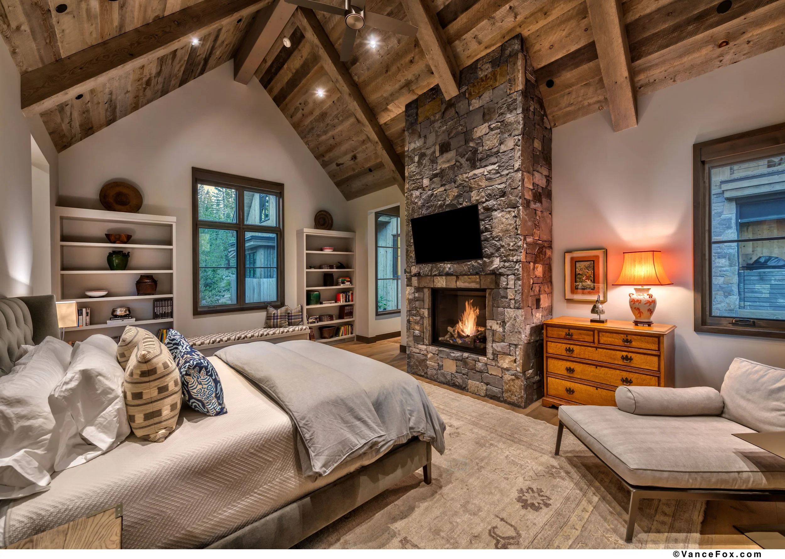 A cozy bedroom with a sloped wooden ceiling, stone fireplace with a mounted TV, and a bed with multiple pillows. There is a wooden dresser with a lamp, an armchair, and built-in white shelves filled with decorative items and books. Windows showcase g