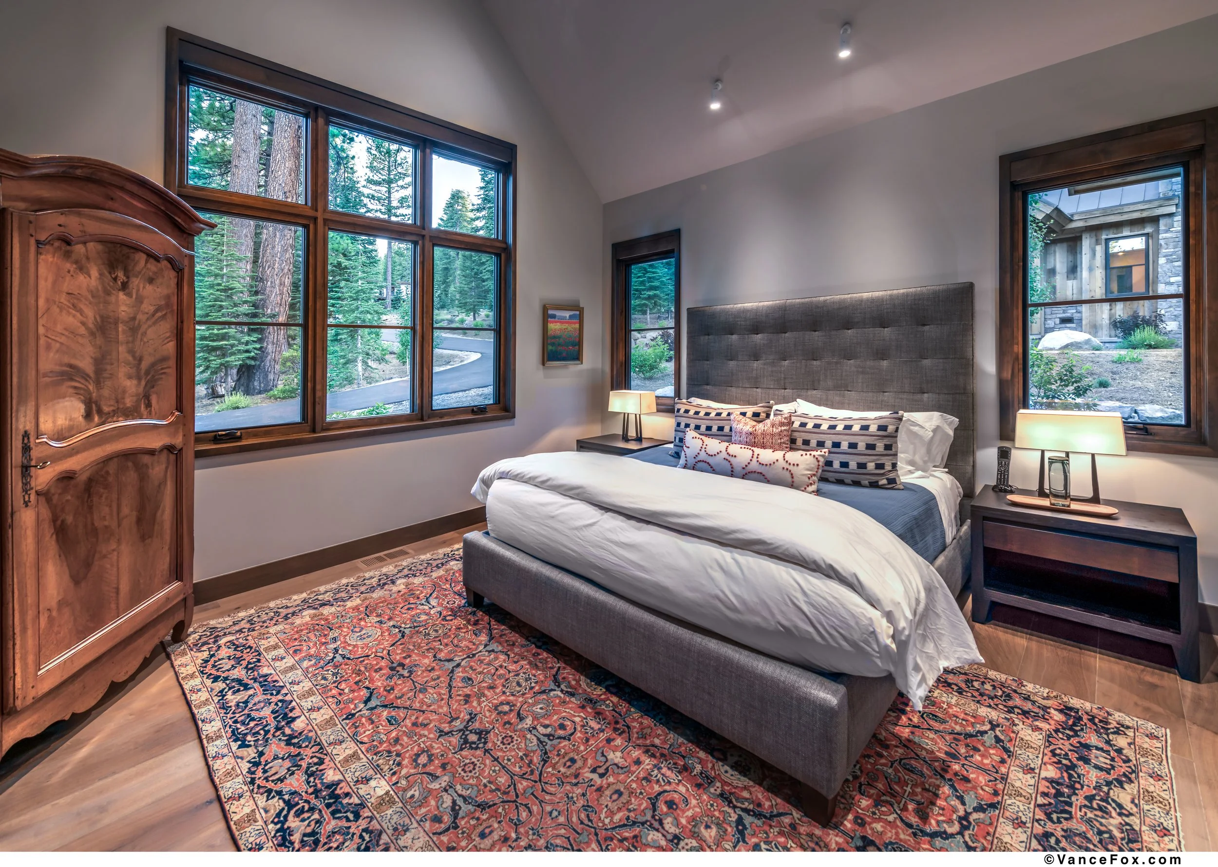 A cozy bedroom with a large bed featuring a tufted gray headboard, decorated with striped and patterned pillows, on a Persian-style rug. There are two nightstands with lamps beside the bed and large windows revealing a lush green forest outside.