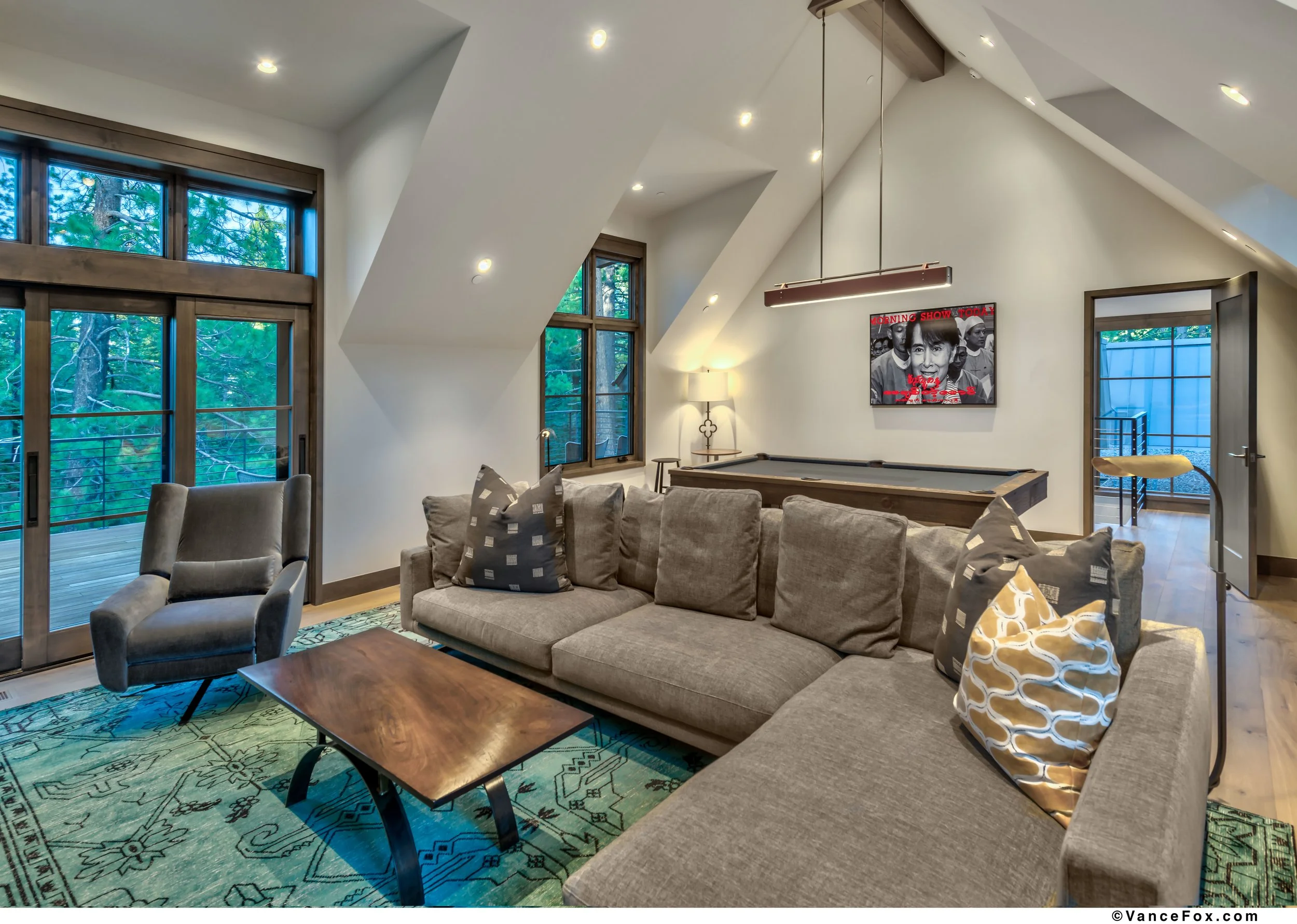 Living room with sectional sofa, patterned pillows, grey armchair, wooden coffee table, patterned area rug, wood-framed glass doors leading to balcony, and windows with trees outside, sloped ceiling with multiple recessed lights, pool table against w