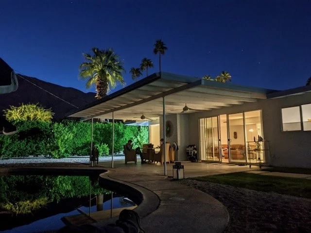Exterior backyard at night with a modern house, outdoor sitting area, pool, palm trees, and a dark sky.