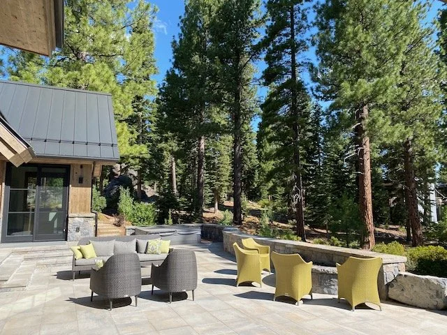 Outdoor patio area with gray and green chairs, a stone wall, and tall pine trees in the background.