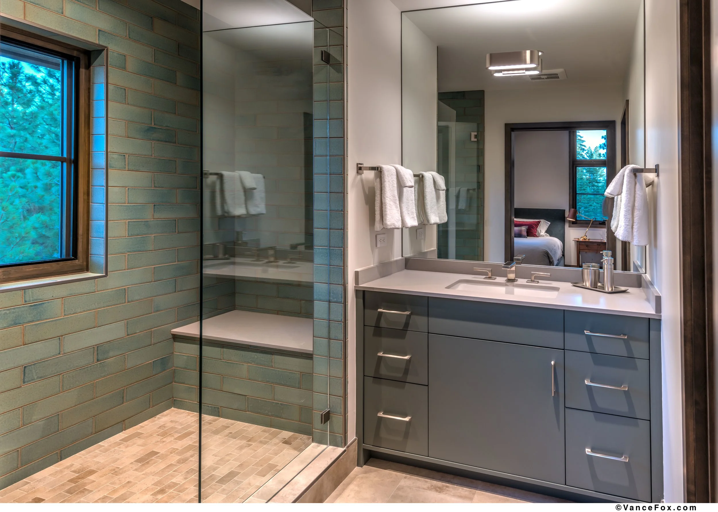 A modern bathroom featuring a walk-in shower with greenish tiles, a large mirror, a gray bathroom vanity with drawers, white towels, a window with a view of trees, and a glimpse of a bedroom with a bed and a nightstand.