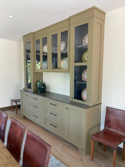 A beige built-in china cabinet with glass doors displaying decorative plates inside, situated in a dining room with wooden chairs and a wooden floor.