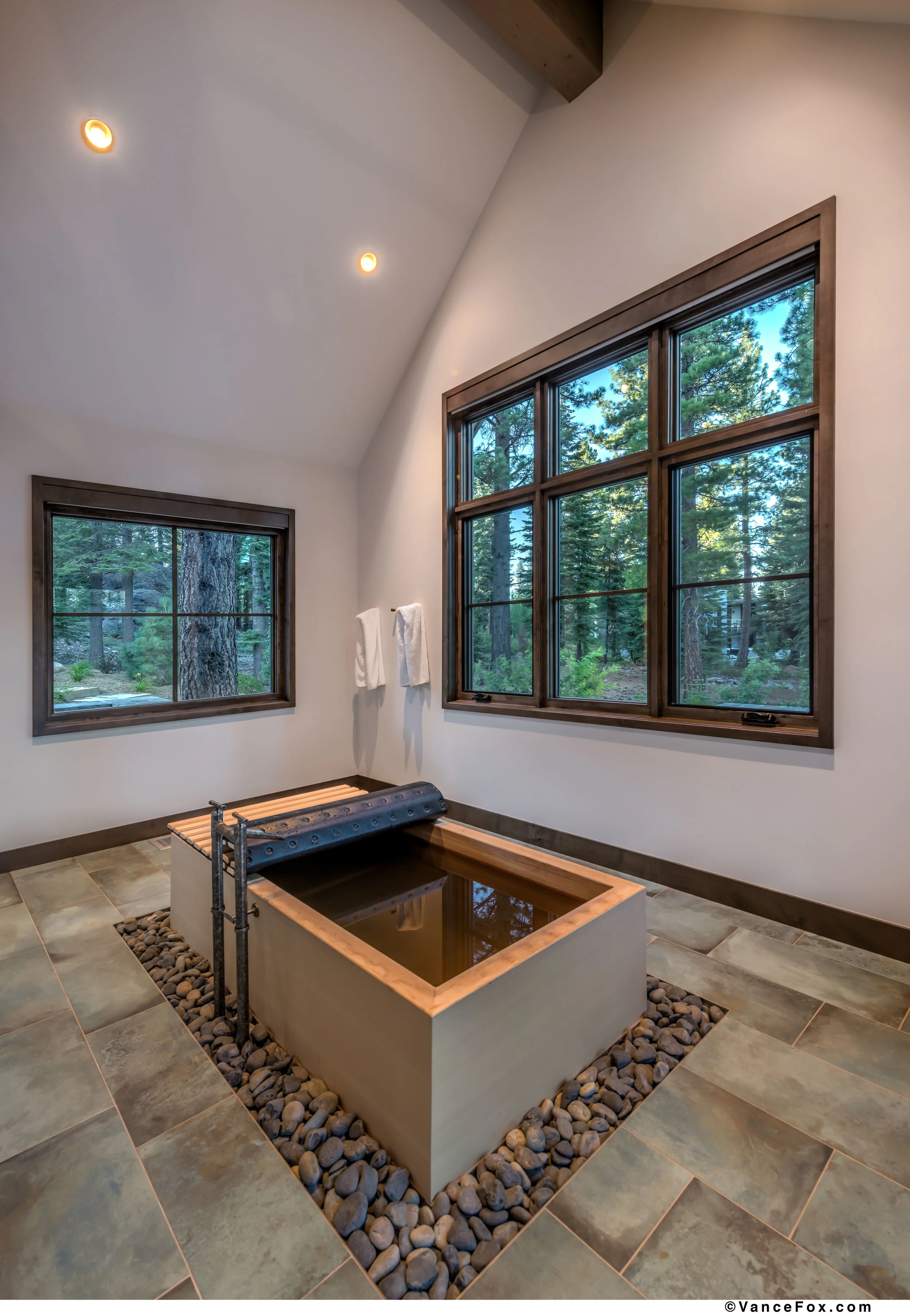 A modern indoor soaking tub surrounded by smooth black stones, with large windows overlooking a forested landscape, wood paneling, and a tiled floor.
