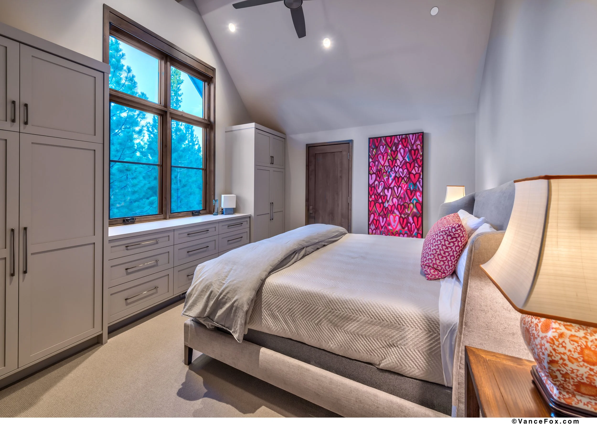 A cozy bedroom with a large bed, gray headboard, white bedding, and pink decorative pillows. There are beige nightstands with lamps, a set of white cabinets and drawers under a large window with wooden trim, and colorful artworks on the wall. The roo